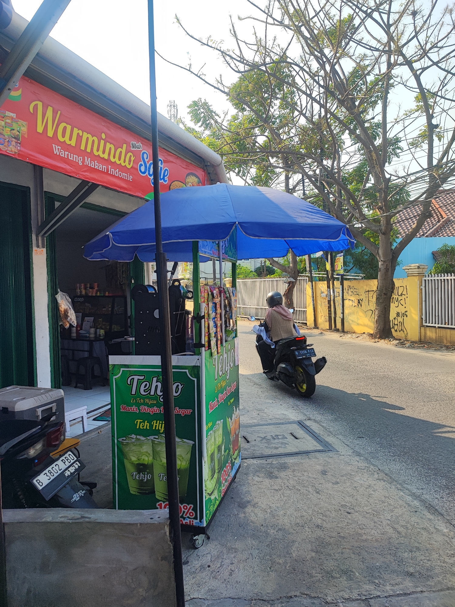 In Satriamekar, Indonesia, a street-side vendor stall is visible on a sunny day. The stall, branded "Warmindo Mbak Sofie," appears to sell instant noodles and beverages. A blue umbrella shades the vendor's cart, which displays promotional images of "Teh Hijau" (green tea). Packaged snacks hang from the cart. To the left of the stall, a parked motorcycle with license plate number B 3802 PBO is partially visible, along with a gray cooler. In the background, a person rides a motorcycle away from the camera down a paved road. The road is lined with a yellow fence, a light blue building, and a large tree with bare branches. Graffiti is present on the yellow wall. The scene depicts a typical urban commercial activity in Indonesia.