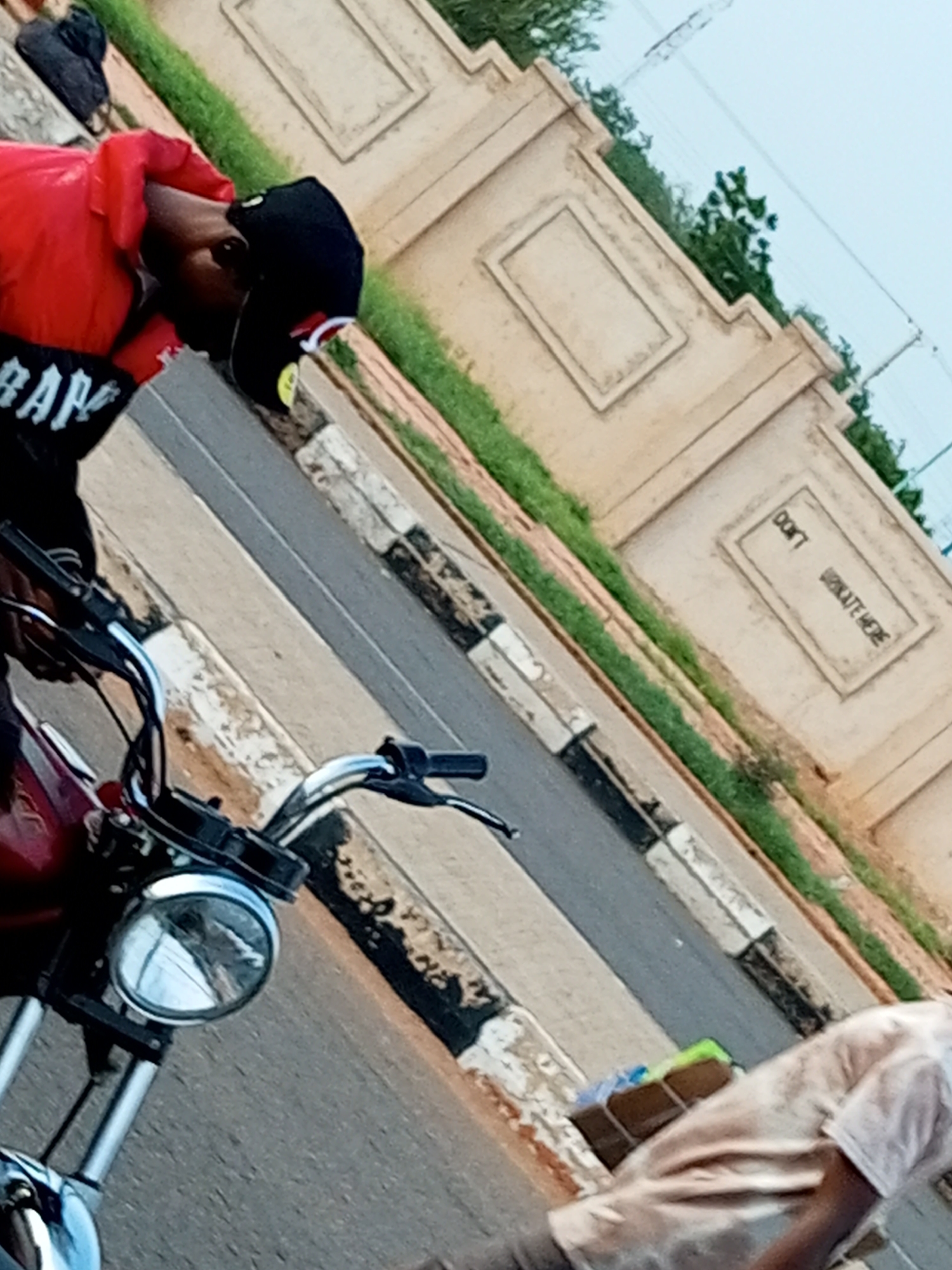 In Gombe, Nigeria, a person wearing a red jacket and a black cap with a yellow emblem is seen leaning over the handlebars of a red motorcycle. The motorcycle's headlight is prominently visible. In the background, a beige wall with decorative rectangular panels runs along a grassy area. On the wall, graffiti in a rectangular box reads "DON'T SKATE HERE". Further in the background, a person in a light-colored shirt is visible near some boxed items. The scene appears to be set beside a road.
