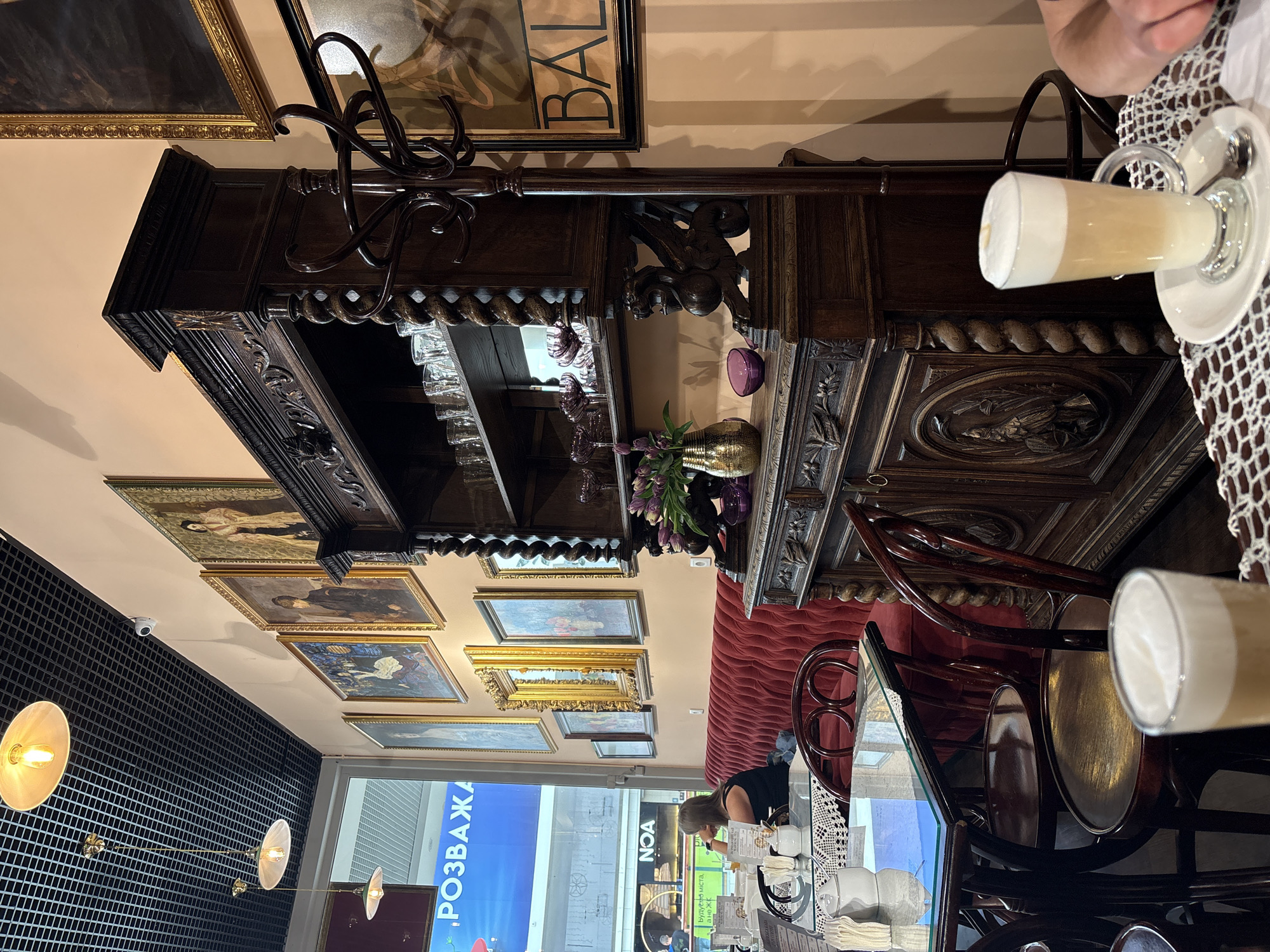 The image depicts the interior of a cafe, likely located in Sokilnyky, Ukraine, given the presence of Cyrillic text on a nearby sign reading "РОЗВАЖА". The cafe has an old-world ambiance with ornate dark wood furniture, including a large display cabinet filled with glasses and decorative items. The walls are adorned with several framed paintings, suggesting an artistic theme. Seating includes a plush red velvet couch and dark wooden chairs with curved backs. A woman is seated on the couch, looking towards a window or display area. On a table in the foreground, a glass of latte with foam art is visible, along with a white ceramic sugar dispenser and a white lacework placemat. The lighting appears to be warm, with decorative pendant lamps hanging from a black grid ceiling.