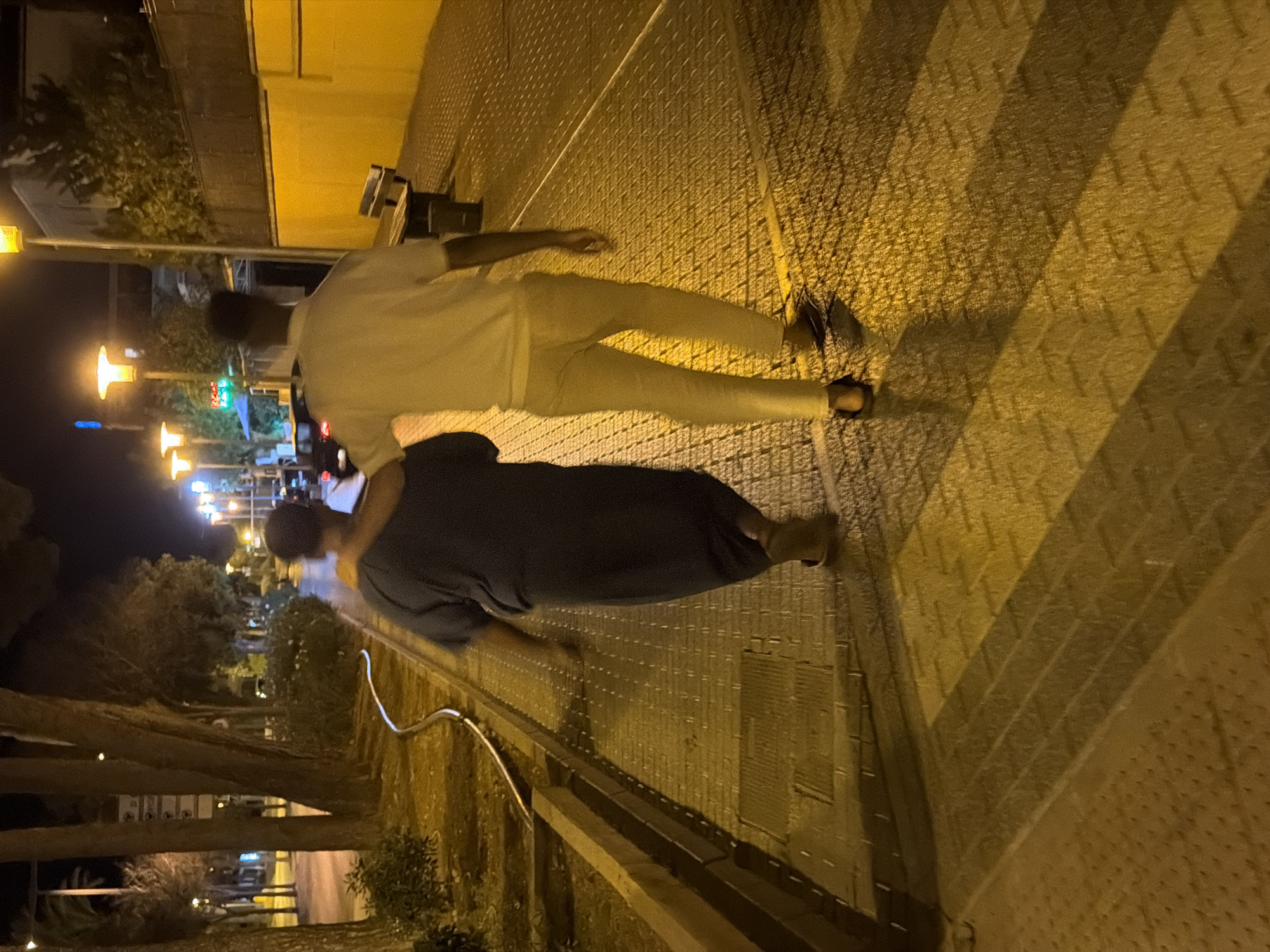 In Calvià, Spain, at night, two individuals walk away from the camera on a pedestrian walkway. The person on the left, wearing a dark, long-sleeved garment, has an arm around the shoulder of the person on the right. The person on the right is wearing a white polo shirt and light-colored trousers. They are walking on a textured, paved surface illuminated by streetlights. Trees line the left side of the walkway, and a yellow building with a bench and trash receptacle is visible on the right. The scene suggests a quiet evening stroll in a Spanish town.