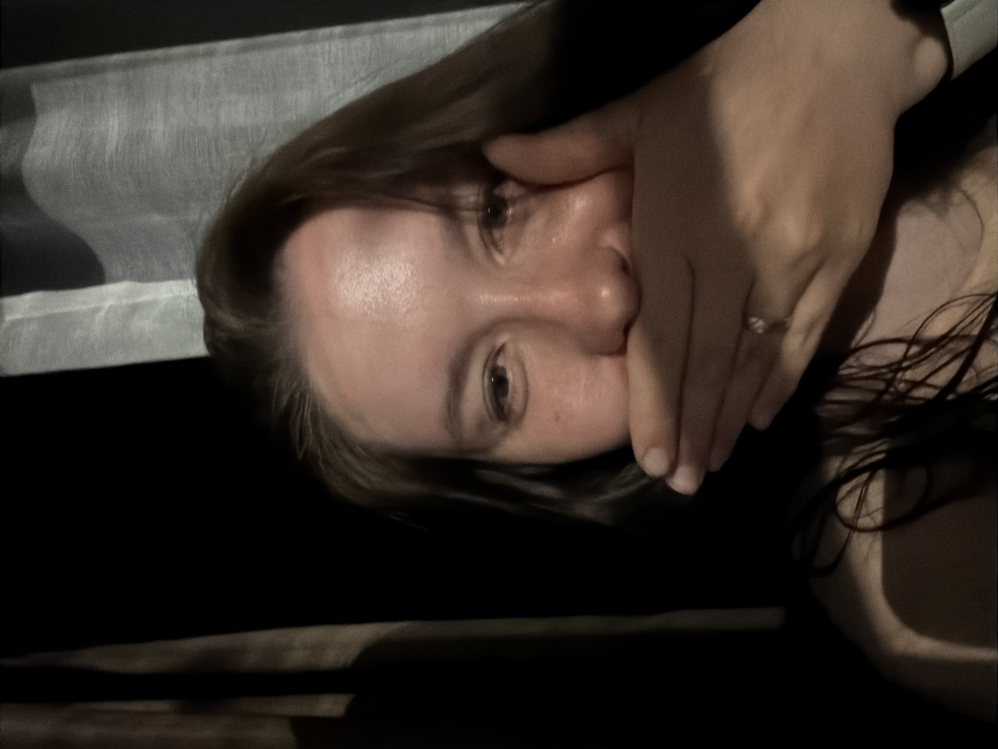 A woman lies on a dark surface, possibly a bed or floor. She is holding her hand over her mouth. The scene is dimly lit. The location cannot be determined based on the image.