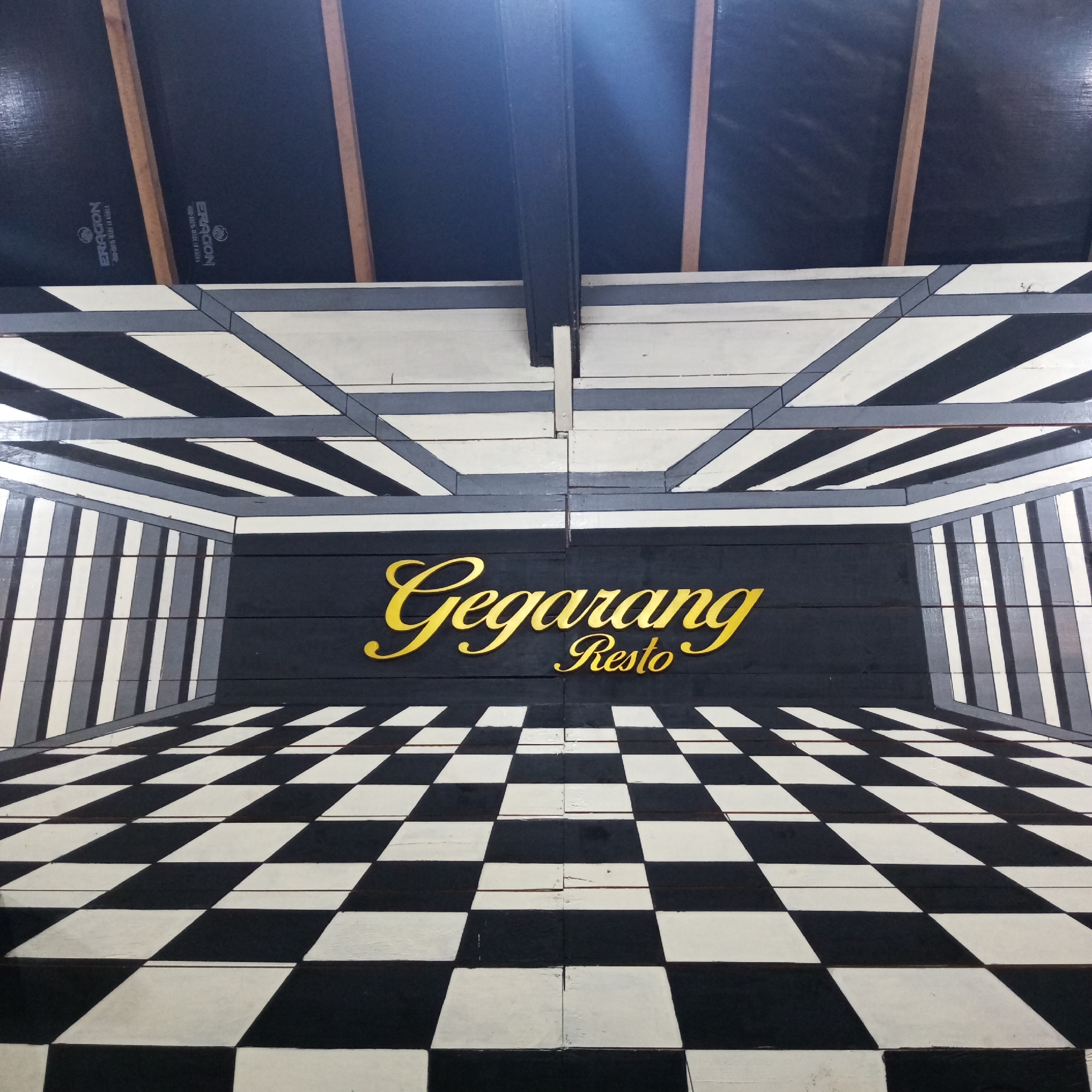 The image shows a mural painted on wooden planks, depicting a checkered floor and walls that create an illusion of depth. The word "Gegararg" is painted in large, gold, cursive lettering, followed by "Resto" in smaller gold lettering below it. The mural is located in Tansaril, Indonesia, likely on an exterior wall or as a decorative element for a restaurant. The mural's perspective makes it appear as if one is looking down a hallway or into a room. The top portion of the image shows wooden beams and black material, possibly part of the building's roofing structure. There are no people or animals visible in the image.