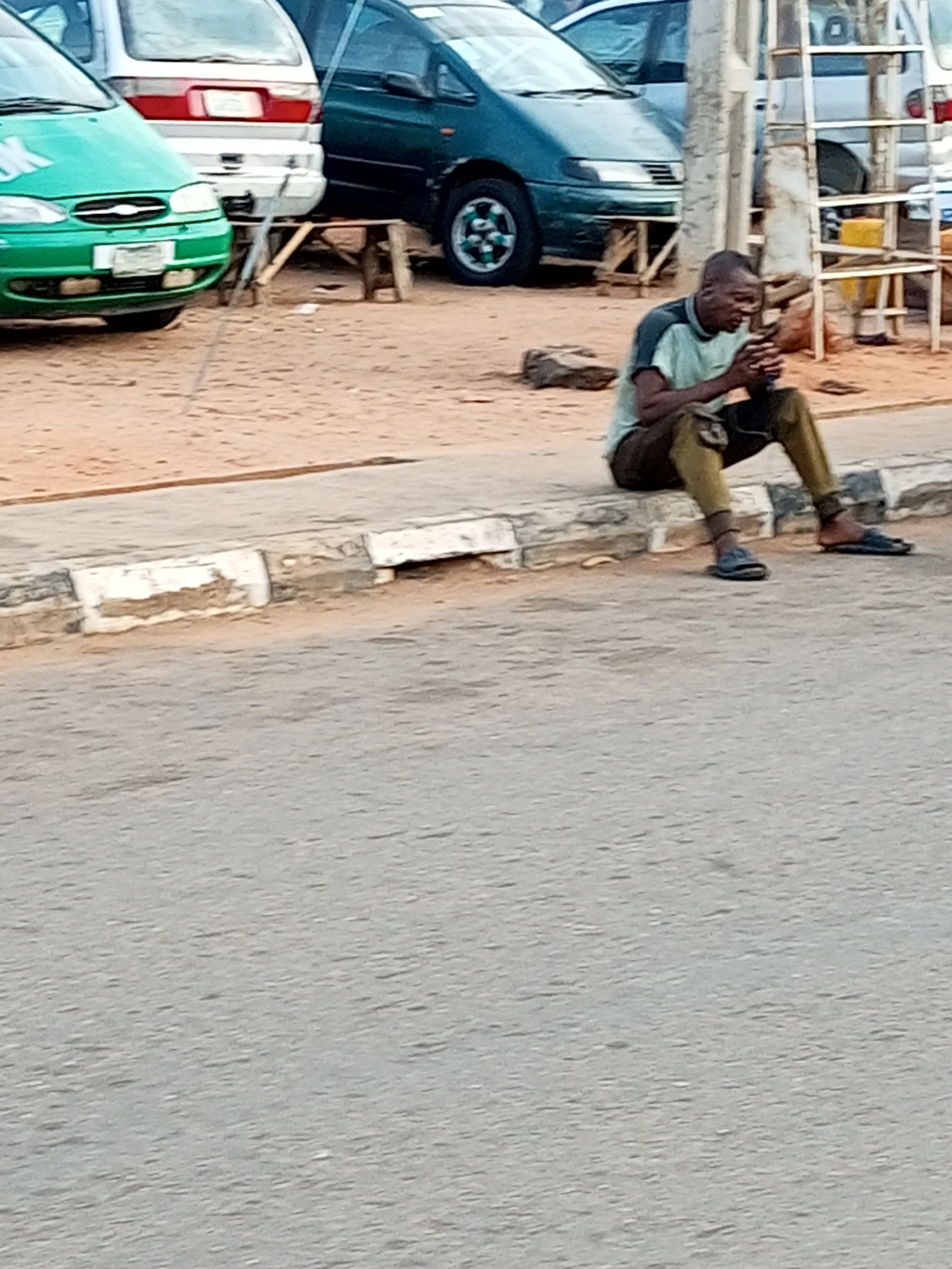 A man of African descent, wearing a light blue t-shirt and olive-green trousers, is seated on a concrete curb. He is holding a mobile phone in his hands and appears to be looking at it. The man is barefoot, wearing dark-colored sandals on his feet. He is positioned in front of a dirt and sand area, with several vehicles visible in the background. To his left is a green car, and behind him, a white van and a dark green van are parked. A wooden structure, possibly a makeshift table or bench, is situated between the green car and the white van. The scene appears to be an outdoor setting in a city, likely Gombe, Nigeria, given the context. The overall environment suggests a roadside or parking area.