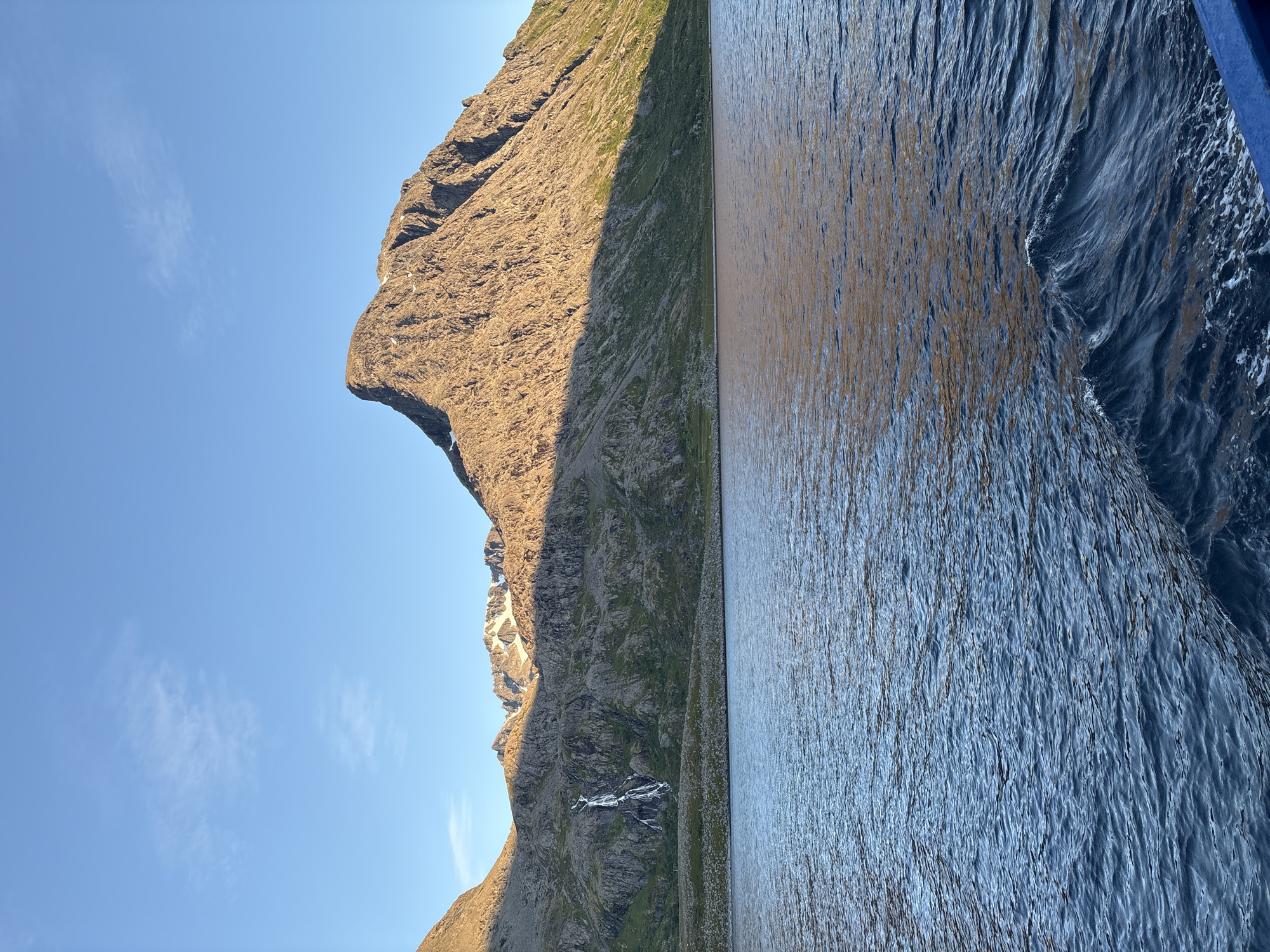 This is a photograph of a coastal mountain landscape taken from a boat. The foreground consists of dark blue water with small waves. In the midground, a rocky, mountainous shoreline is visible, with a waterfall cascading down the side of a hill on the left. The mountains are partially illuminated by sunlight, casting shadows. The peaks in the distance are snow-capped. The sky is clear and blue with a few wispy clouds. The scene is likely captured during a sunset or sunrise, given the lighting. The location is Arnøyhamn, Norway.