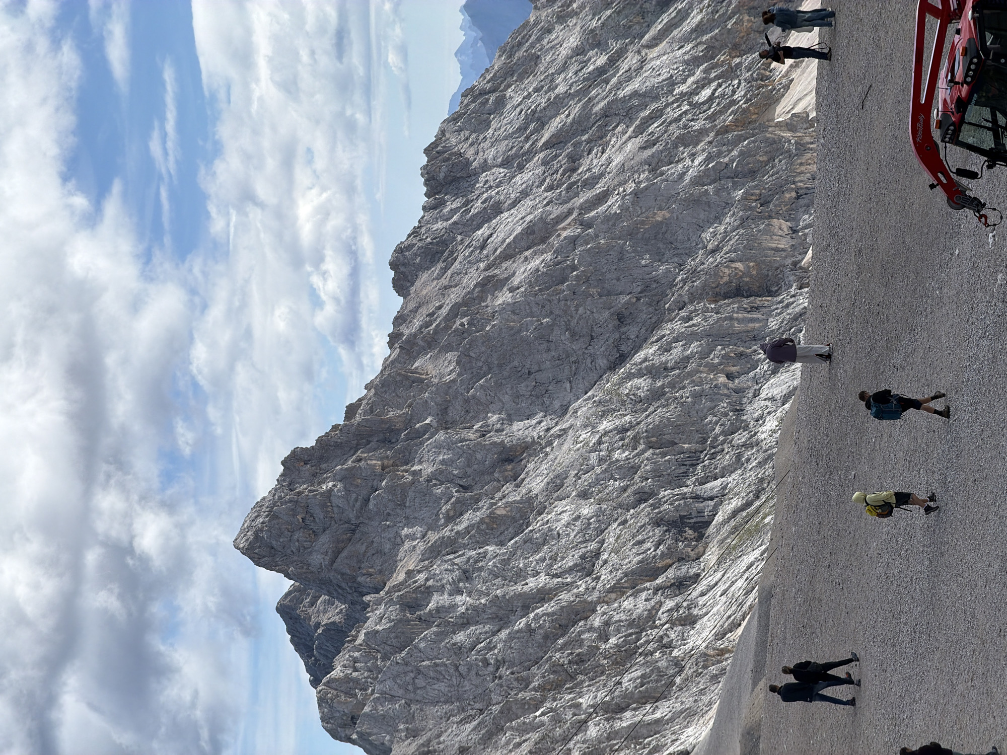 The image depicts a mountainous landscape with several people on a gravelly plateau in front of a large, rugged rock face. The sky is partly cloudy with a mix of blue sky and white clouds. In the foreground, a red snow groomer or similar vehicle is partially visible on the right. Several individuals are scattered across the plateau, some walking, some standing. On the left, a couple walks away from the camera. Further into the scene, a person in a yellow hooded jacket walks towards the right, followed by another person with a blue backpack. On the far right, a group of three people stands together, with one appearing to take a photo or video. The rock face dominates the background, showcasing its rough texture and steep incline. The location is the Zugspitze massif near Garmisch-Partenkirchen, Germany, likely at the viewing platform area accessible by cable car or cogwheel train.