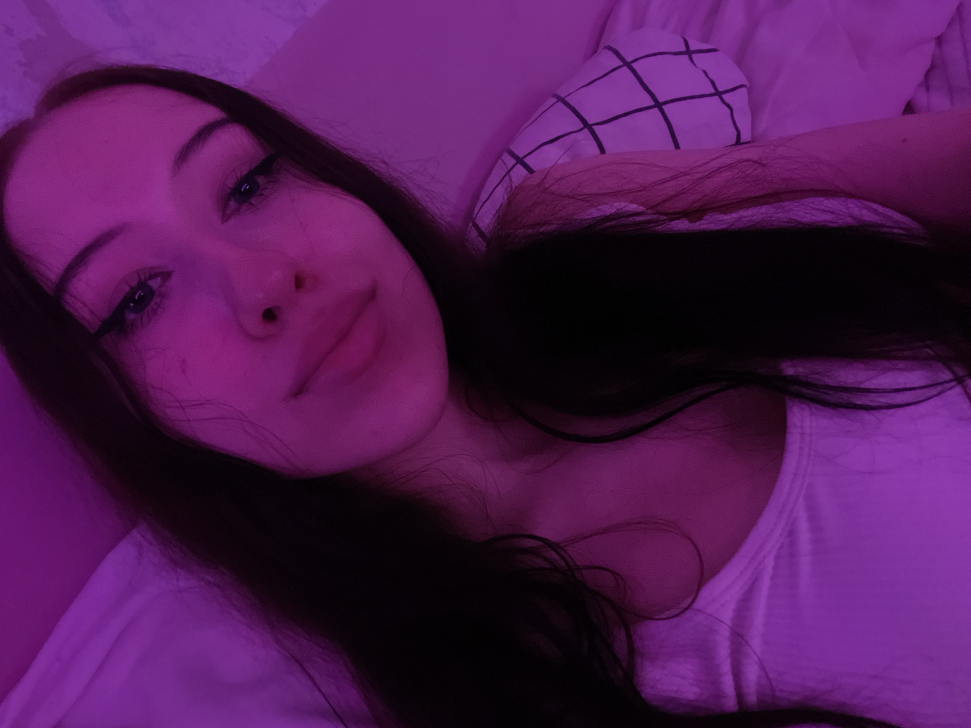 The image shows a close-up, selfie-style shot of a young woman with long, dark hair and makeup. She is wearing a white tank top. The scene is lit with purple lighting, giving the image a distinct, moody atmosphere. There is a pillow with a black grid pattern visible in the background. The overall setting appears to be indoors, possibly a bedroom. The provided hints suggest details about her face, hair, neck, and shoulder area, as well as the tank top and the pillow. There is no information that could identify the specific location within Votkinsk, Russia.