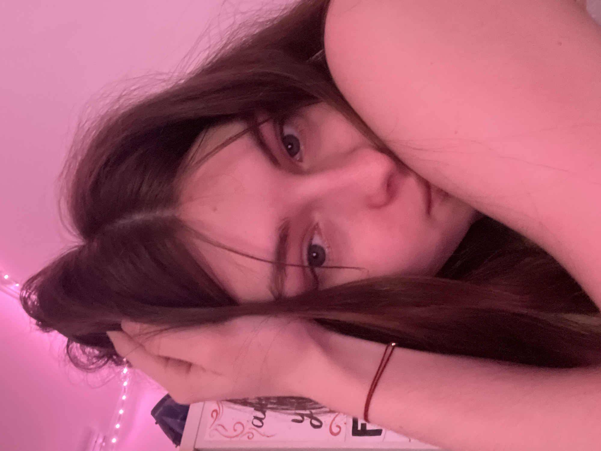 A young woman lies on her side in a room illuminated by pink lighting. Her dark brown hair is partially obscuring her face. Her left hand is near her face, and a red string is visible on her left wrist. There are decorative lights visible in the lower-left corner. The background includes a patterned surface with red and white detailing. No location information from Saint Petersburg, Russia is discernible.