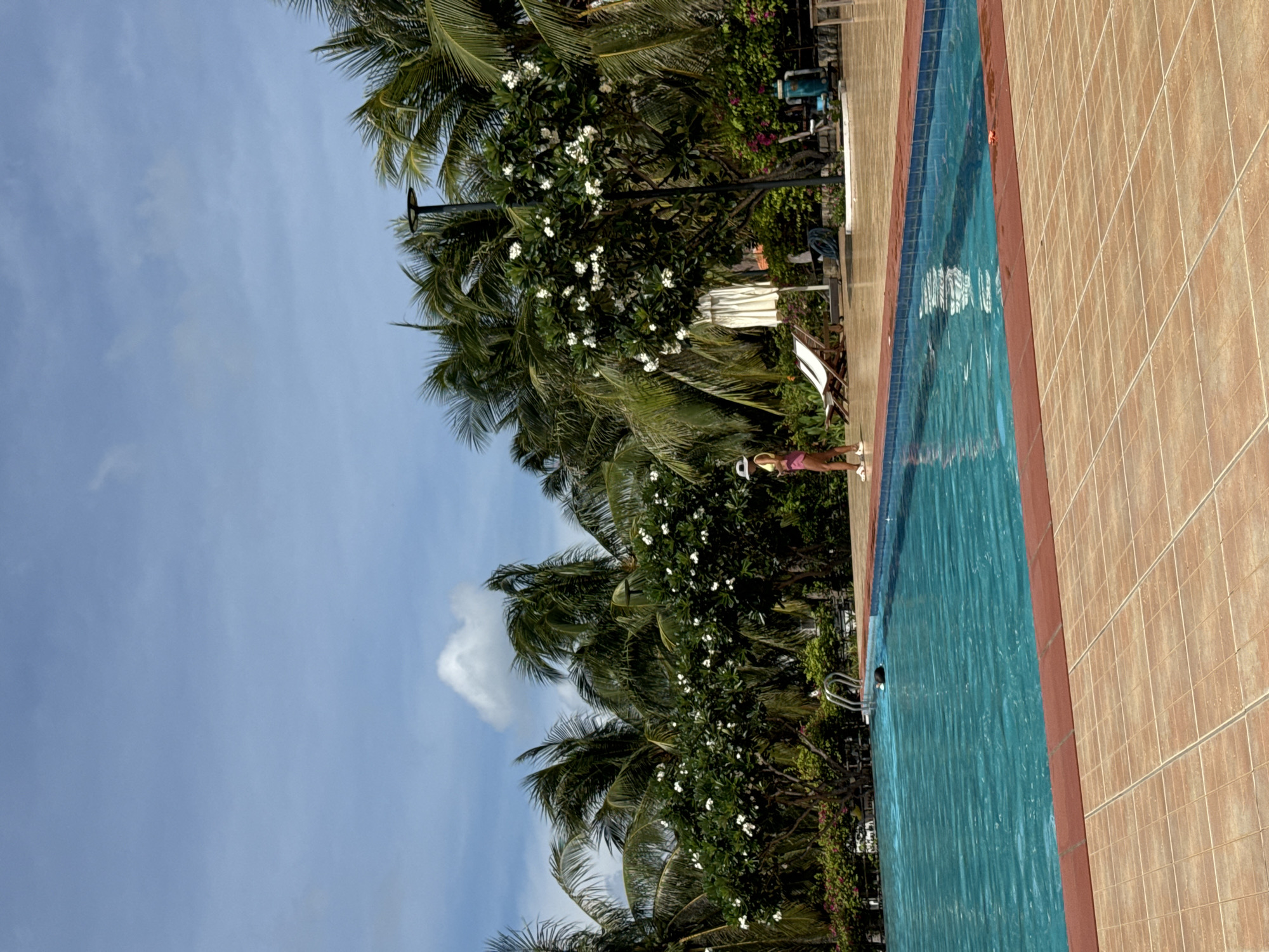 The image depicts a resort swimming pool area in Phu Thuy Ward, Vietnam. A woman in a yellow top and pink shorts stands on the tiled poolside, wearing a white hat. To her left, two empty lounge chairs are placed on the tiled ground. The turquoise water of the swimming pool fills the foreground, with a set of silver pool stairs visible on the left side of the pool. In the water, a black object, possibly a flotation device or an animal, is partially submerged. The background is lush with palm trees and flowering bushes, suggesting a tropical environment. A modern lamppost stands near the center background, and a white umbrella is visible behind the lounge chairs. The sky is a clear blue with scattered white clouds.