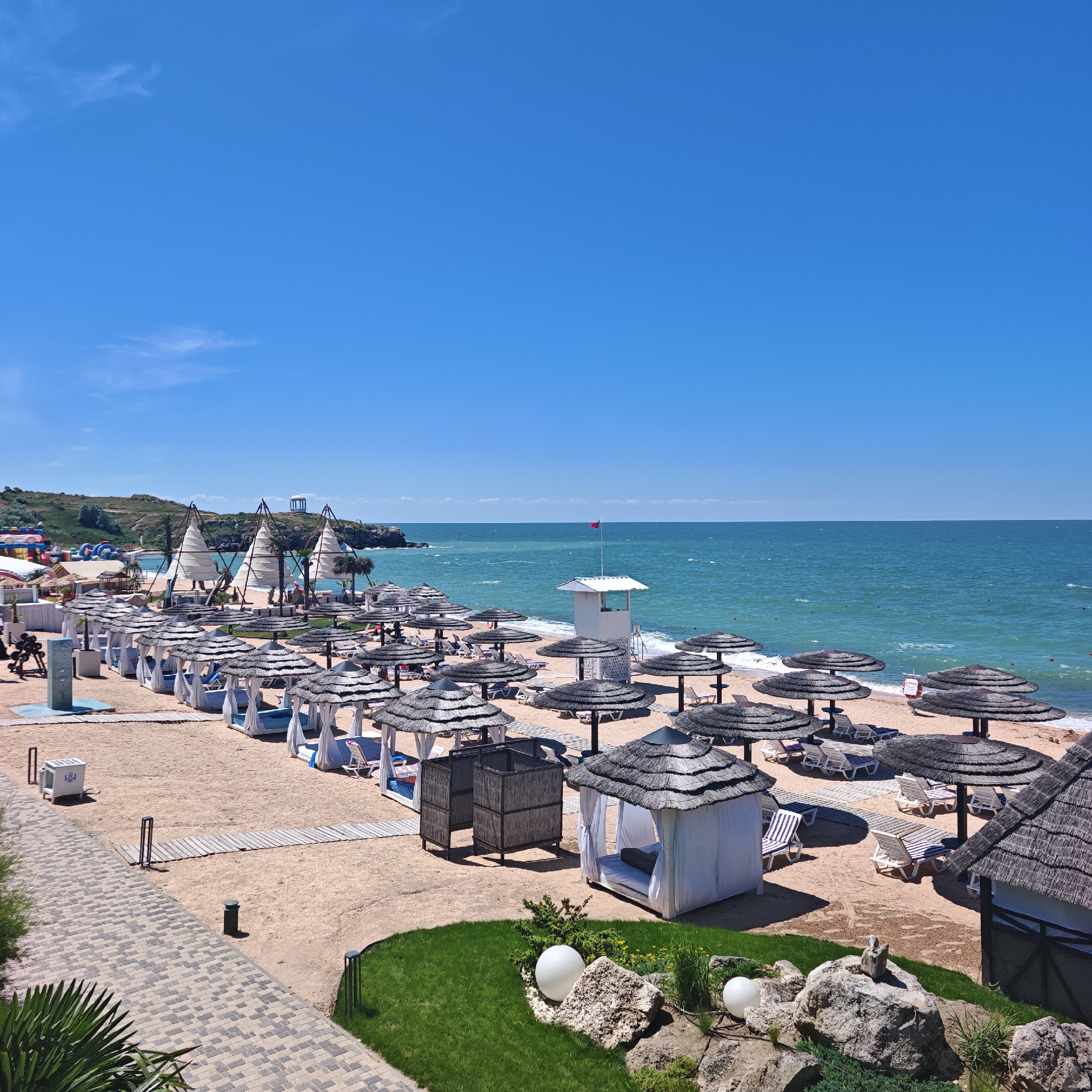 This is a wide-angle, eye-level outdoor photograph taken on a sunny day at a beach resort. The scene depicts a well-maintained beach with numerous amenities for visitors. The foreground features a paved walkway made of grey and beige interlocking pavers, bordered by lush green grass and decorative rocks, one of which holds two white sphere-shaped lights. To the left of the walkway, a large palm frond is visible.
The beach itself is covered in light-colored sand. A significant portion of the frame is occupied by rows of beach cabanas with white curtains and thatched roofs, as well as many large, dark-colored umbrellas providing shade over lounge chairs. Some lounge chairs are occupied by people, though they are not clearly discernible. In the middle ground, there are several unique, conical-shaped structures with white fabric sails, resembling small tents or shelters. Further back, inflatable structures, possibly for children's entertainment, are visible on the beach.
The sea is a vibrant turquoise color and appears calm with gentle waves lapping at the shore. A lifeguard tower, a white structure with a red flag flying from its top, stands near the water's edge. In the distance, a rocky coastline with some greenery and a white rotunda-like structure on a hill is visible. The sky is a clear, bright blue with a few wispy clouds. The overall impression is a resort destination prepared for a busy tourist season. The location is identified as Мысовское сельское поселение, Ukraine.