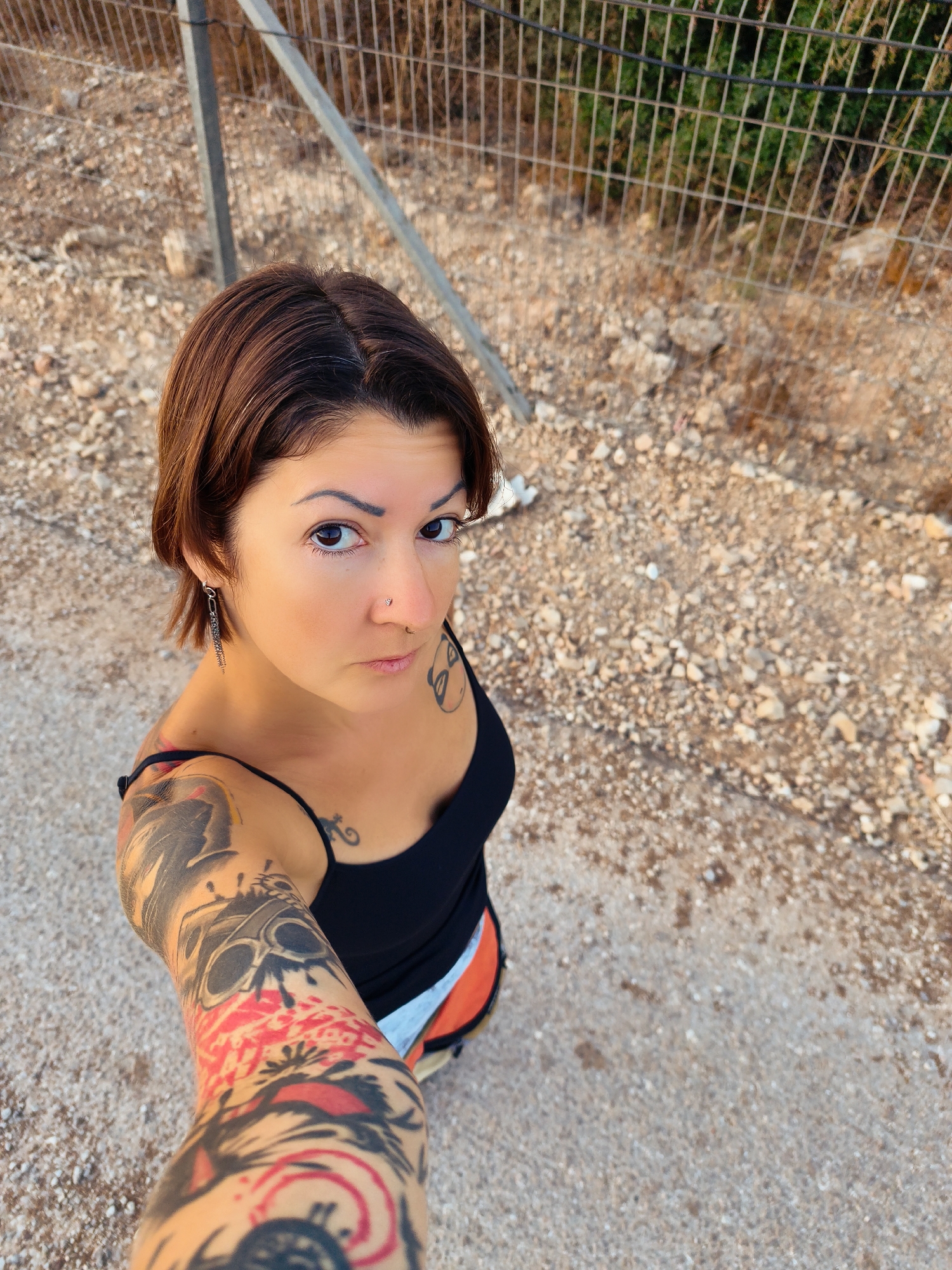 A selfie of a tattooed woman standing outdoors. She has short brown hair, dark eyes, and a nose ring. She is wearing a black tank top and orange shorts. Her left arm is covered in tattoos, including a skull. The background is a gravelly area with a chain-link fence and some dry vegetation. The image appears to have been taken in the city of Lachish Regional Council, Israel, given the dry, rocky terrain and the presence of a chain-link fence, which is common in such areas.