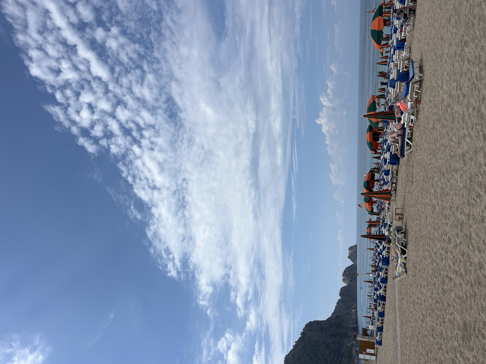 The image captures a beach scene in Alanya, Turkey, identifiable by its characteristic cliffs and coastal features. A line of beach umbrellas and sun loungers stretches along the sandy shoreline, with people reclining and relaxing. The umbrellas are mostly orange and green, adding color to the scene. The sea is visible in the distance, with a rocky outcrop rising from the water. The sky is predominantly blue, with patches of clouds. In the foreground, the beach sand takes up a significant portion of the image. A mountain is located at the left side of the image.
