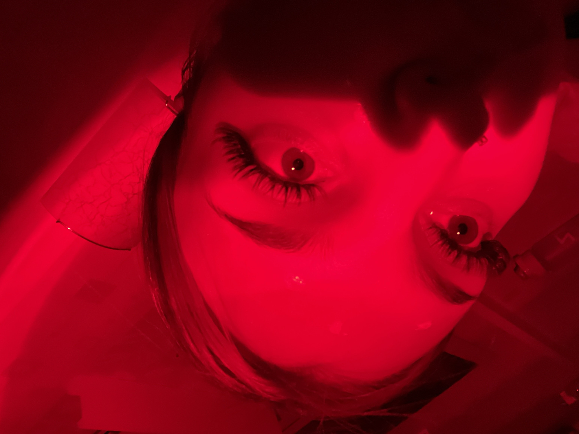 The image presents a close-up, high-angle portrait of a person, likely female, bathed entirely in a striking, monochromatic red light. The subject's face is oriented with the chin towards the top of the frame and the forehead towards the bottom, looking upwards into the camera. Both eyes are prominently visible, featuring long, dark eyelashes, dilated pupils, and bright white reflections on the irises.
The intense red illumination creates deep shadows, particularly obscuring the nose and mouth area, adding to the dramatic and mysterious atmosphere. Dark hair frames the face on both sides, blending into the red glow of the background. On the left side of the frame, near the subject's ear, a small, thin object, possibly a wire or an accessory, is visible. Further to the left, a rectangular object with a textured surface, perhaps paper or fabric, is partially illuminated by the red light.
The scene suggests a low-light environment intentionally lit by a strong, artificial red light source. No specific activity is discernible beyond the posed portrait. The overall mood is intense and somewhat eerie, emphasizing the eyes as the focal point. There is no visible text in the image. The location is unknown.