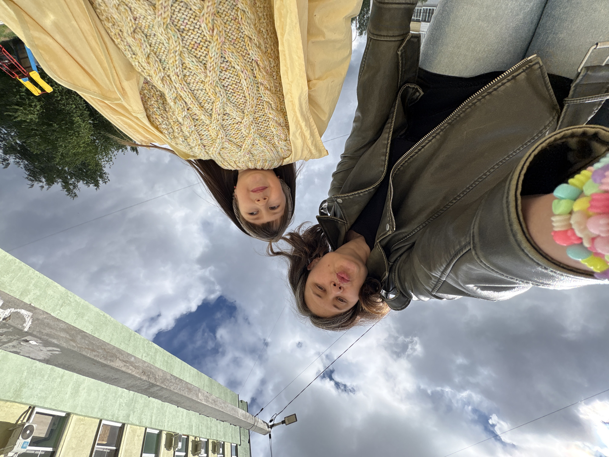 An upward-angle shot captures two young women looking down at the camera, set against a cloudy daytime sky. The scene is likely outdoors in Chaikovsky, Russia.
The woman on the left has long, dark hair and is wearing a yellow knitted sweater with a cable pattern over a light yellow garment. She also has a headband and small pink earrings. The woman on the right has shoulder-length, dark brown hair with some lighter highlights, and is dressed in a dark olive-green zippered jacket over a black top, paired with light-wash jeans. She has a multi-colored pastel beaded bracelet on her right wrist and is playfully making a "duck face."
In the background, a partly cloudy sky dominates. Below the horizon, a light green building with several windows and an air conditioning unit is visible. A prominent concrete utility pole with an attached street light cuts across the lower part of the frame. To the far left, green foliage and part of a red and yellow structure, possibly a playground, can be seen. The weather appears overcast but clear, suggesting a daytime shot. No visible text is legible in the image.