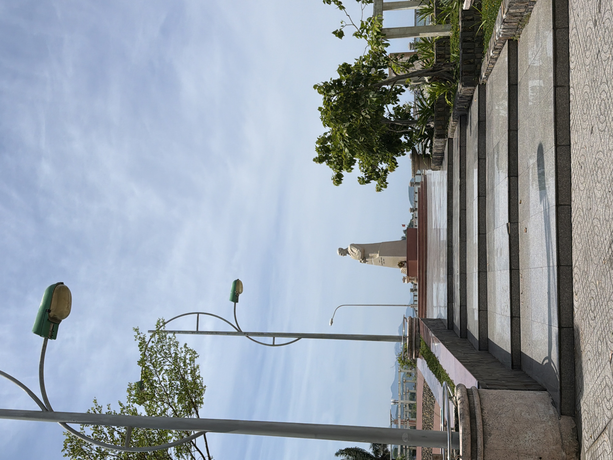 The image captures a monument in Nha Trang Ward, Vietnam. A statue stands atop a tall pedestal on a raised platform, which is accessible by a wide set of stone steps. The steps are made of polished stone, reflecting the sky. Flanking the steps are stone planters and manicured greenery. Two street lamps, one with a green housing, are visible on the left side of the frame, with a large tree also present. In the background, a body of water and distant mountains can be seen under a partly cloudy sky. The overall scene is serene and suggests a public memorial or landmark.