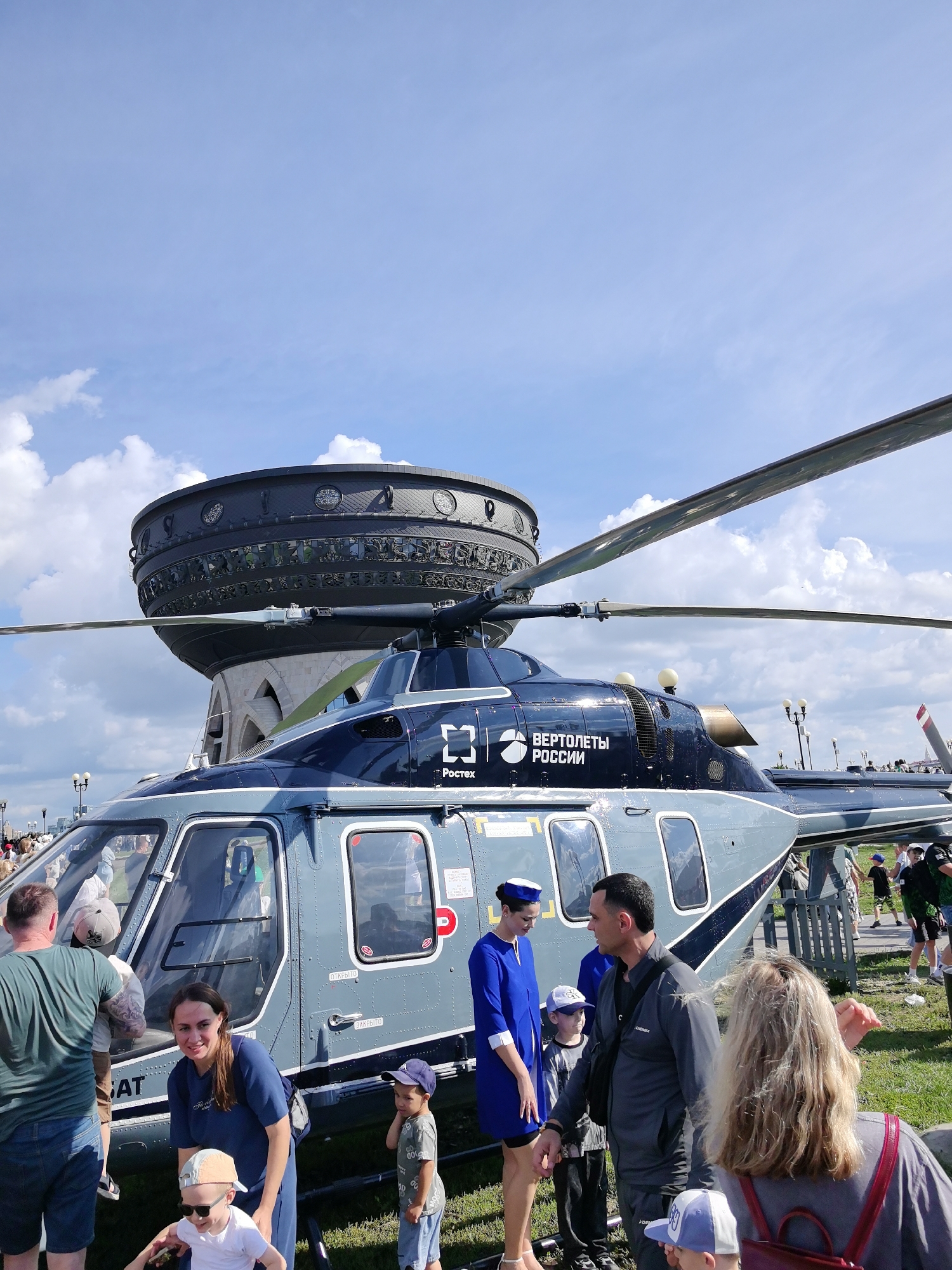 An outdoor daytime scene in Kazan, Russia, features a large, grey and dark blue helicopter with "ВЕРТОЛЕТЫ РОССИИ" (Russian Helicopters) and Rostec logos visible. Several people are gathered around the helicopter, including adults and children. A woman in a blue uniform, possibly a flight attendant, stands near the helicopter's door. In the background, a distinctive circular structure with ornate detailing and windows is visible, which is part of the "Kazan Family Center" or "Kremlevskaya Embankment". The sky is blue with scattered white clouds.