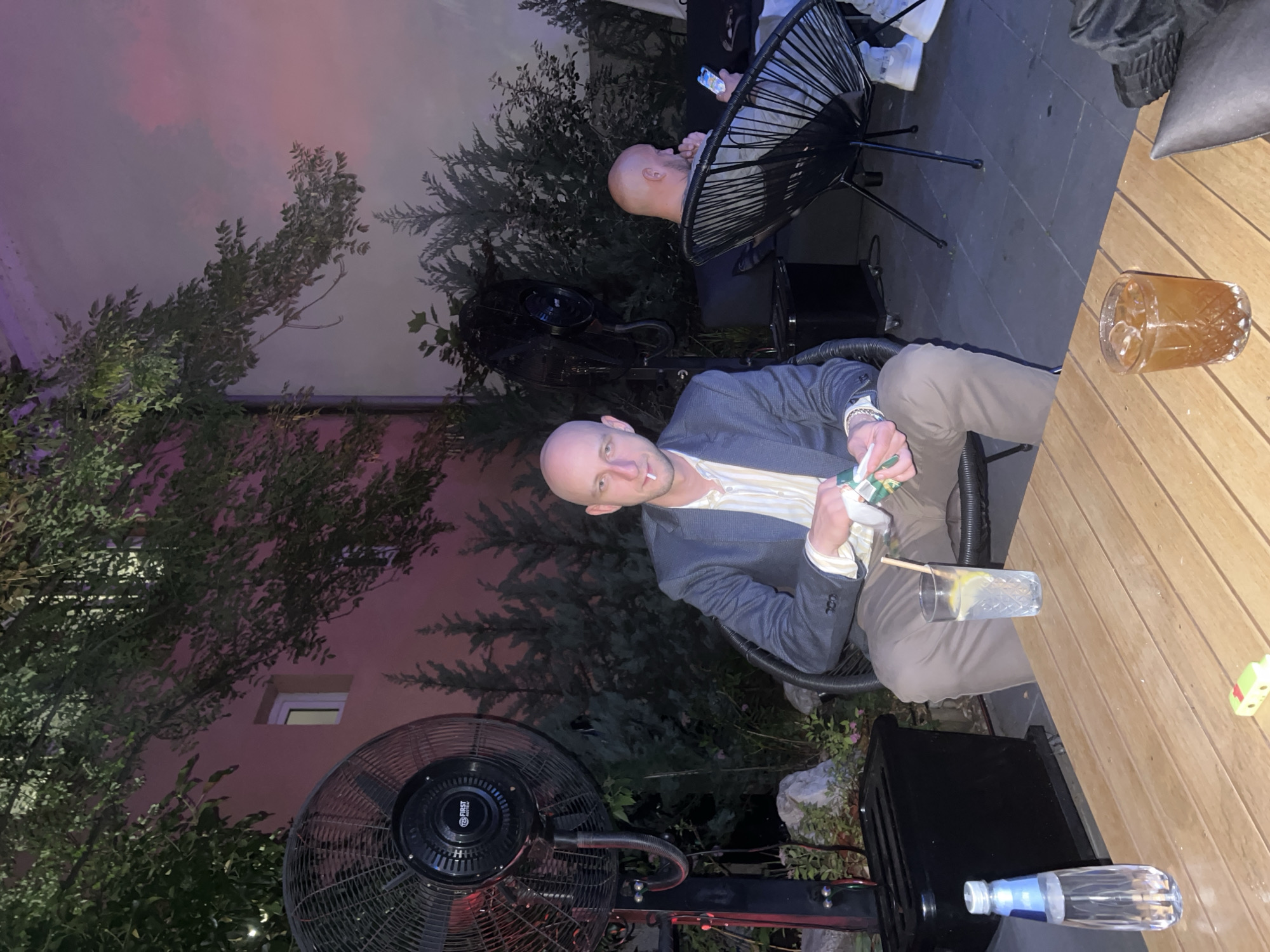 This outdoor evening image, taken in Sofia, Bulgaria, features two men on a paved patio surrounded by vegetation and a building wall. The primary subject, a bald man with light stubble, is seated in a dark wicker chair on the left, looking directly at the camera. He wears a blue-grey blazer over a light-colored striped shirt and tan trousers, with a cigarette or roll-up in his mouth and a packet of tobacco or cigarettes in his left hand. On the wooden table in front of him are a clear glass with ice, a lemon slice, and a straw, a plastic bottle of water, and a small green object.
To the right, a second bald man in a grey t-shirt is seated in a matching wicker chair, facing away from the camera and engaging with a smartphone. His white sneakers are visible. Also on the wooden table is a textured glass containing an amber liquid with ice.
The environment includes a large black industrial fan, branded "FIRST Austria," on the left, with red illumination at its base. The background consists of dense green foliage, including various bushes and trees, set against a building wall illuminated with soft pink and purple light, revealing a small white-framed window. A black utility box or container is positioned next to the primary subject's chair.