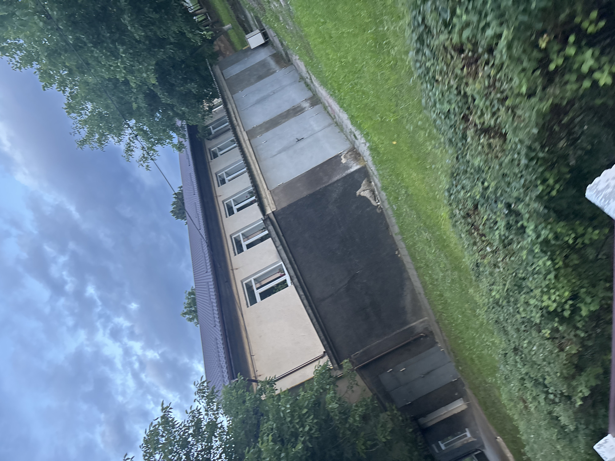The image shows a building with multiple garages. The building appears to be two stories tall, with windows visible on the upper floor. The garages are aligned in a row, each with a separate door. The building is situated in an outdoor environment with a grassy lawn and trees. The sky above is cloudy, suggesting the possibility of rain or overcast weather. The perspective of the image is slightly tilted, giving a dynamic angle to the scene. There are no people visible in the image, and no specific actions are depicted. The overall scene conveys a sense of an everyday urban or suburban setting, possibly in the early evening or a gloomy day.