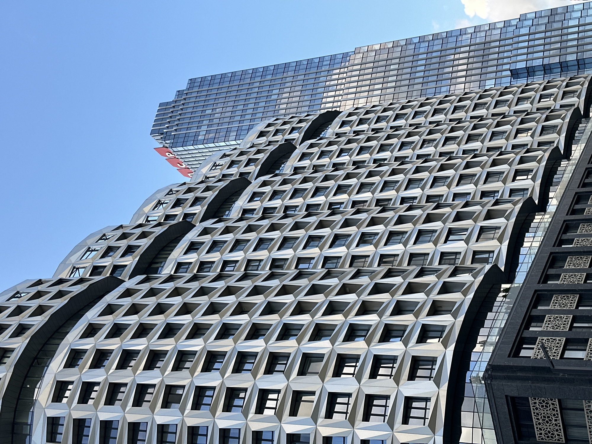 The image depicts a low-angle view of modern architecture in Moscow, Russia, under a clear blue sky. In the foreground, a curved building with a distinctive facade featuring a repeating geometric pattern of recessed window units dominates the left side of the frame. The facade appears to be made of a light-colored, reflective material. Below this structure, a dark-colored building with a glass railing is visible.
To the right and behind the foreground building, a tall, slender skyscraper rises into the sky. This skyscraper has a facade composed of numerous glass panels, reflecting the surrounding environment. Prominently displayed at the top of the skyscraper are large red letters forming the logo "ICP." A few wispy white clouds are visible in the upper right corner of the sky. The scene captures a contemporary urban landscape with striking architectural designs.