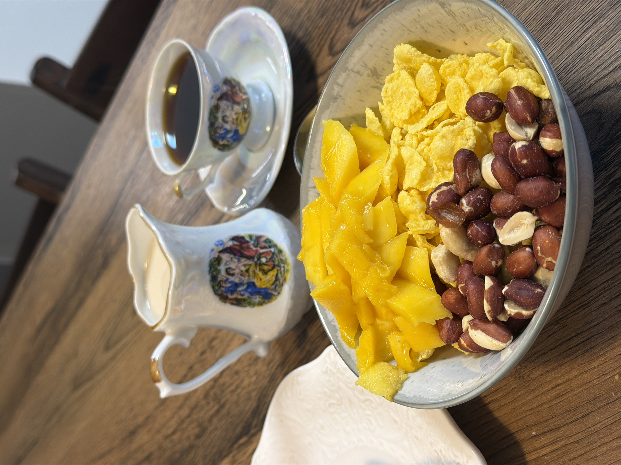A close-up, eye-level shot captures a breakfast spread on a wooden table. In the foreground, a light blue-grey bowl contains a mix of ingredients: chopped mango pieces, cornflakes, and peanuts. To the left of the bowl, a white cloth napkin is visible. Behind the bowl, a decorative white creamer jug with a gold rim is filled with milk, and next to it, a white teacup and saucer hold black coffee. The background shows the backs of two dark wooden chairs. There are no people visible in the image. The scene is set indoors on a wooden surface. The image does not contain any information that would allow for its location to be identified in the city of Perm, Russia.