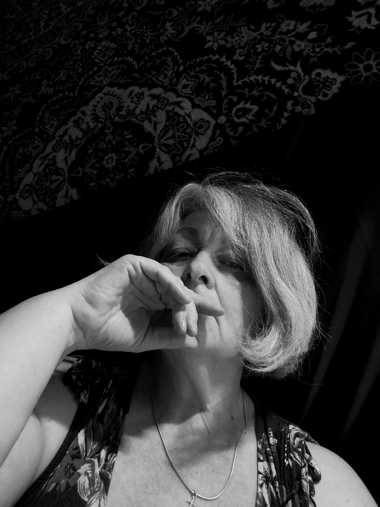 A black and white, close-up, high-angle, medium shot captures a woman from the shoulders up in a dimly lit room. The woman, with short, light-colored hair, is looking down and to the right, with her right hand covering her mouth and nose. She is wearing a floral print top and a silver chain necklace with a cross pendant. The background consists of a dark, patterned fabric, possibly a rug or tapestry, and a dark curtain. The scene appears to be indoors, with dramatic lighting casting shadows across the woman's face and the background. There is no visible indication of the city of Krasnodar, Russia.
