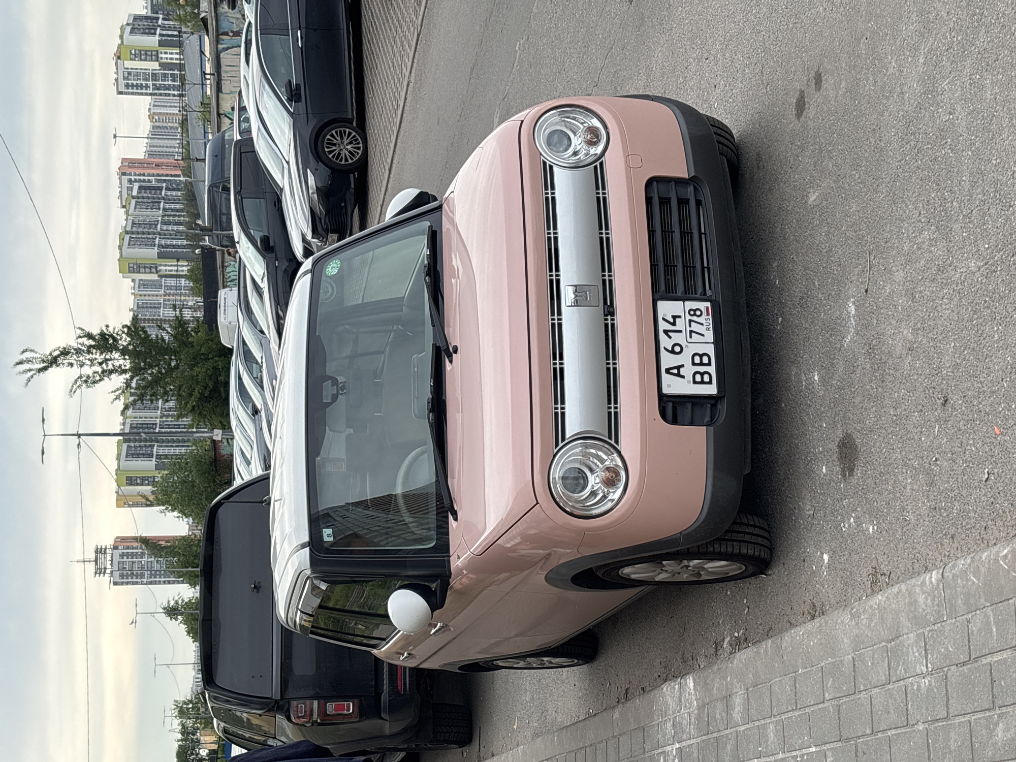 An eye-level shot captures a vibrant pink car, likely a Suzuki Alto Lapin, parked on an asphalt street with a concrete block sidewalk to its right. The car, in the foreground, faces slightly towards the right, showcasing its distinctive rounded headlights and a silver grille with a small emblem. Its front license plate, visible on the lower left, reads "A 614 BB 778 RUS".
Behind the pink car, a line of other vehicles, primarily dark-colored SUVs or vans, are parked. In the background, a modern residential complex stretches across the horizon, featuring numerous high-rise apartment buildings with varied facades in white, grey, green, and hints of orange. Trees are sparsely distributed between the parked cars and the distant buildings, and several streetlights are visible.
The scene appears to be an urban or suburban street during the daytime, under an overcast or partly cloudy sky, indicating diffused light. No people are visible in the frame. The overall atmosphere is mundane, depicting a typical parking situation.
