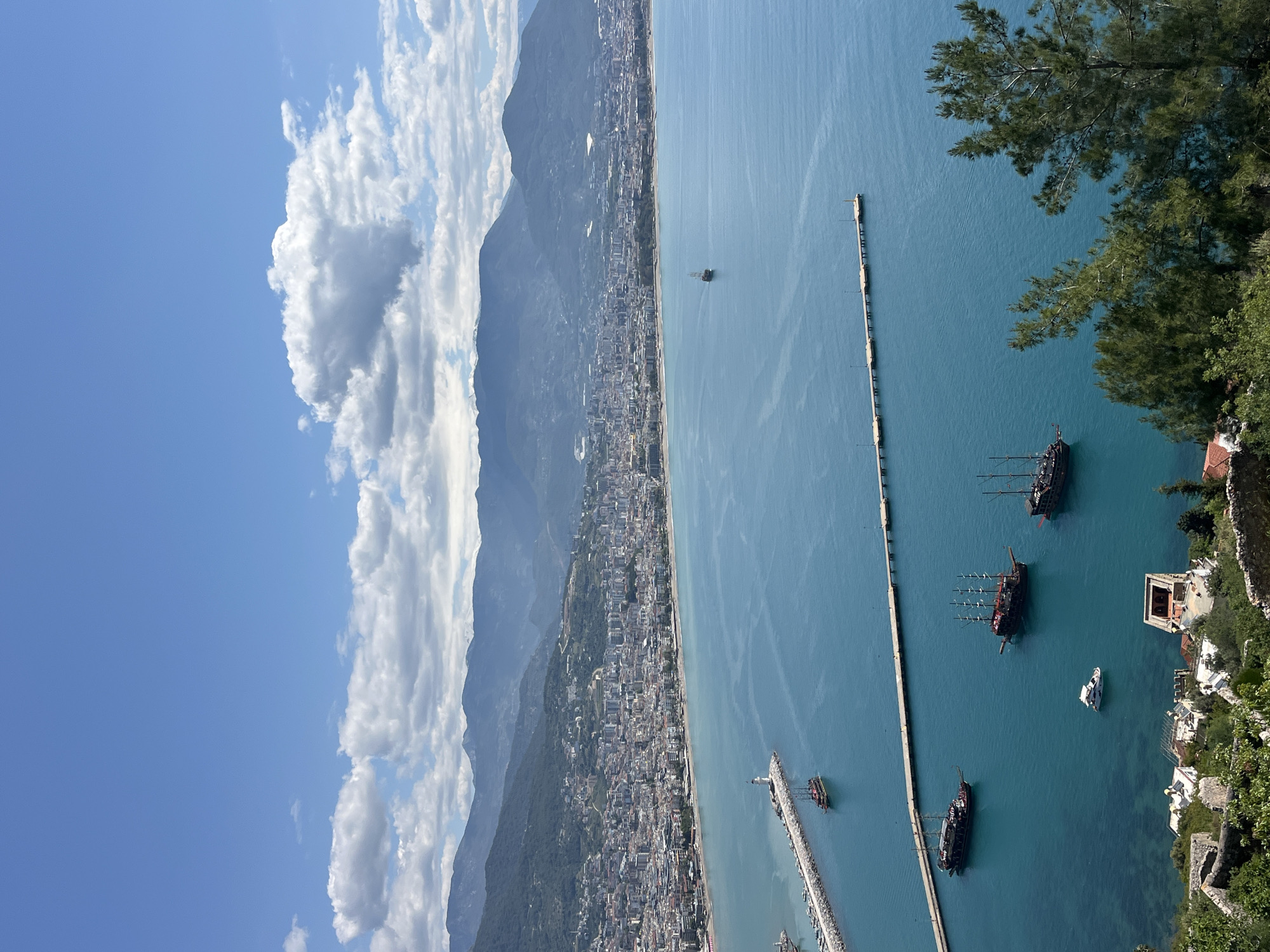 The image captures a high-angle view of Alanya, Turkey, focusing on the coastline and harbor. The clear turquoise water is dotted with several pirate-themed tourist boats. Two long piers extend into the sea from the city’s edge. Mountains and the urban sprawl of Alanya are visible along the coast. The sky is bright blue with cumulus clouds. The view is taken from a vantage point where trees and structures are visible.