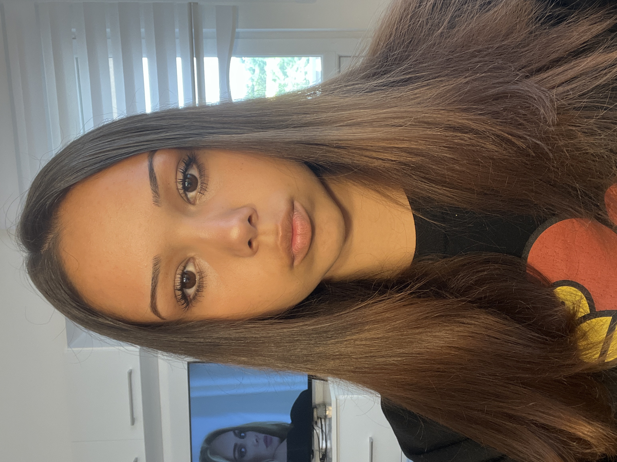 A young woman with light brown skin and long brown hair poses for a selfie in an indoor setting, likely a home. Her face is the focal point, with a neutral expression and soft lighting. Behind her, vertical blinds cover a window. To her left is a television screen, displaying another woman. The location is Unknown, Unknown.