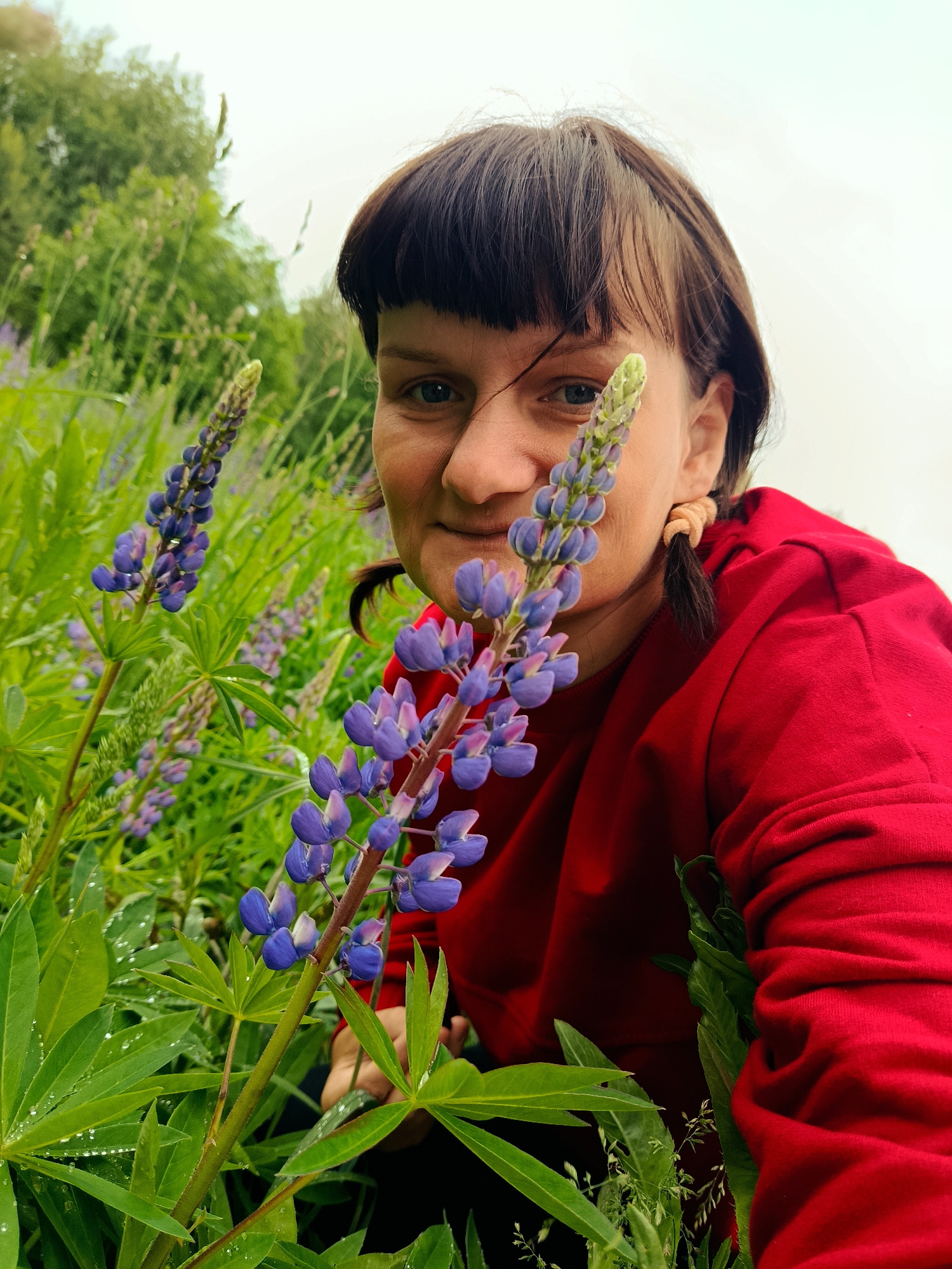 The image displays an outdoor scene in Большая Уса, Russia, featuring a woman amidst dense green foliage and flowering lupines under an overcast sky. The woman, positioned in the center-right, has medium-brown hair with bangs and visible pigtails secured by light-colored hair ties. Her light-colored eyes are directed towards the viewer, and her lips form a slight smile. She is wearing a red long-sleeved garment, identifiable by its fabric texture and folds. A prominent stalk of purple and blue lupine flowers, with a lighter green undeveloped tip, stands in the foreground, partially obscuring the woman's face. To the left, another fully bloomed lupine stalk is visible further back. Numerous other lupine flowers and extensive green leaves, many bearing clear water droplets, fill the surrounding environment. The background is softly blurred, showing additional green vegetation and a bright, featureless sky. The perspective is low, placing the viewer at eye level with the flowers and the lower portion of the woman's body.