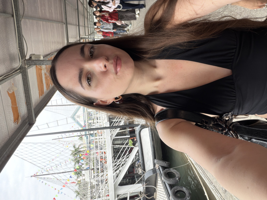 A woman takes a selfie on a boat. She wears a black v-neck top. A black purse hangs from her shoulder. The boat has a tall mast adorned with colorful flags. Other boats and water are visible. People stand on a walkway in the background. A structure, possibly a covered walkway, is overhead.