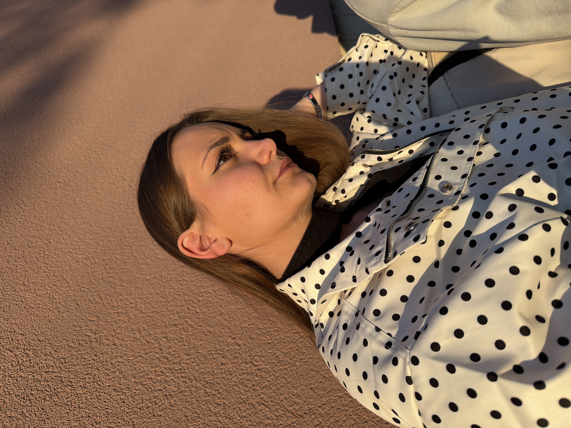 A young woman lies on a brown, textured surface, possibly concrete. She is wearing a white jacket with black polka dots over a black top. Part of a light-colored object, possibly a piece of furniture, is visible in the upper right corner. A shadow falls across the surface in the upper left corner. She has light brown hair and is wearing a watch on her left wrist. The location is unknown, and there is no confirmation that this photo was taken in Buhovo, Bulgaria.