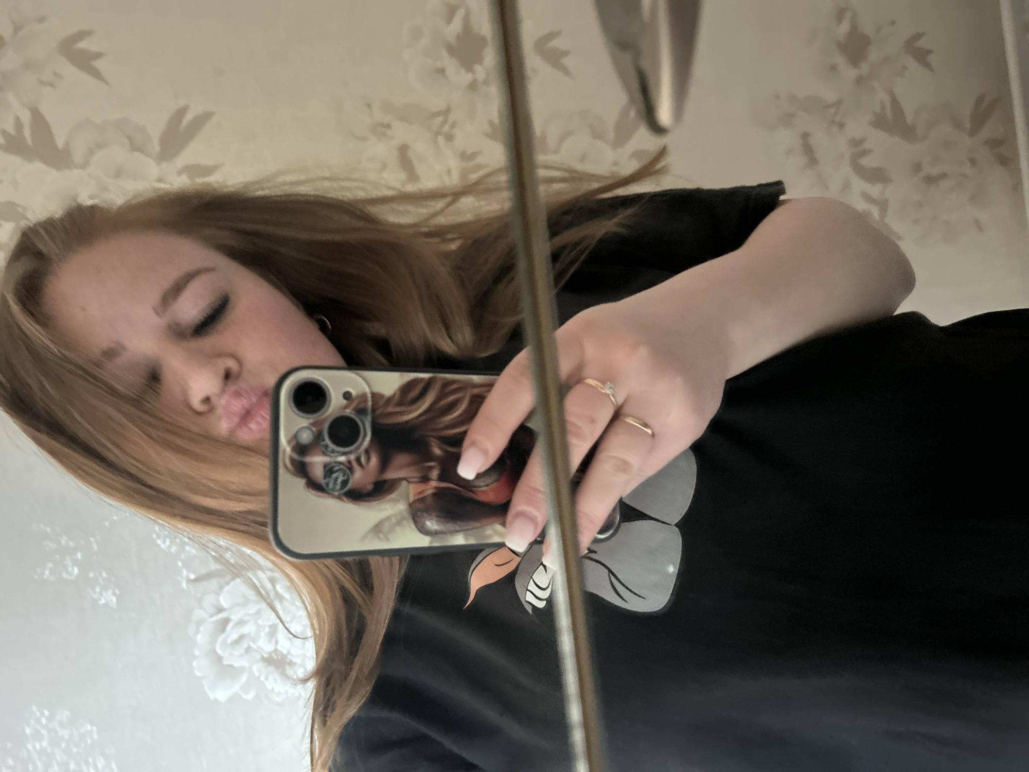The image shows a woman taking a selfie in a mirror. She is holding a smartphone with a case featuring an image of a woman with sunglasses and a flower. The woman in the image has light brown hair, is wearing a black t-shirt, and has rings on her fingers. The background shows a floral patterned wallpaper and a portion of what appears to be a bathroom fixture. No information is present in the image that could lead to locating this scene in the city of Chaikovsky, Russia.