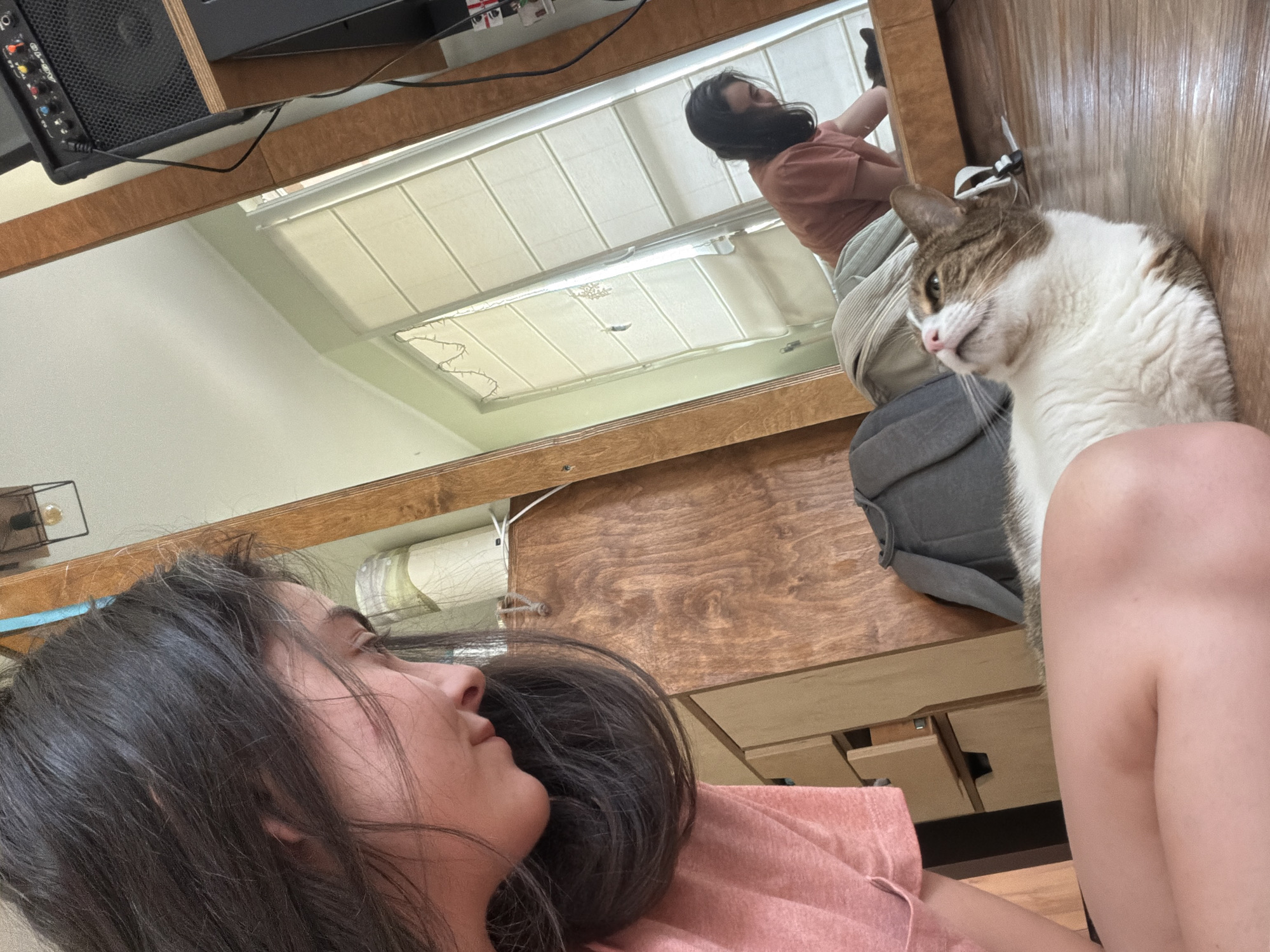 In an indoor setting with wooden floors and wooden furniture, a young woman with dark hair is crouched down, looking at a cat. The woman is wearing a pink t-shirt. The cat, which is white with brown and gray tabby markings, is lying on the floor, looking up at the woman. A large mirror in the background reflects the scene, showing the woman and the cat from a different angle. Part of a music amplifier is visible on a shelf above the mirror. The environment appears to be a room in a home.