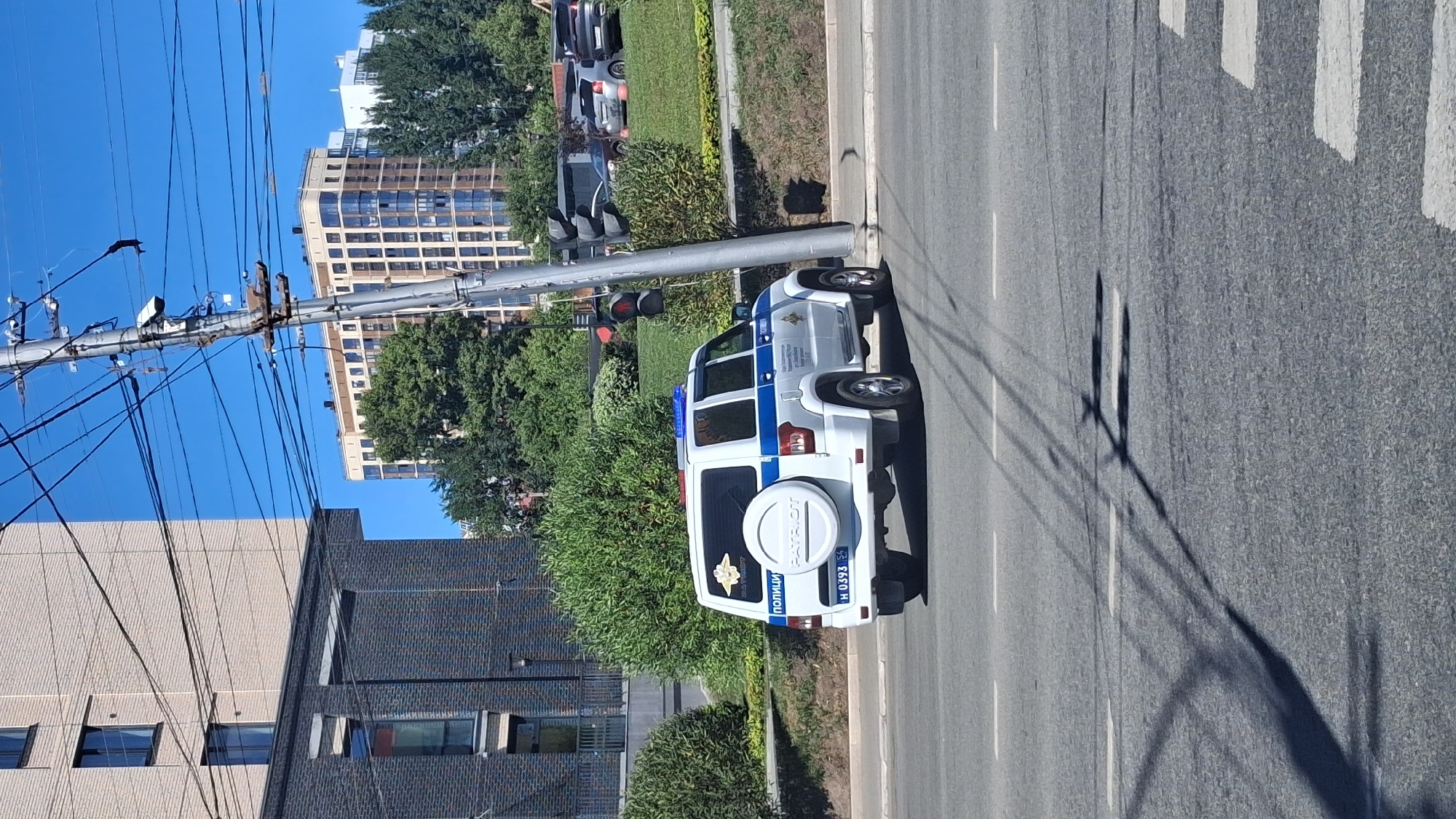 In Novosibirsk, Russia, a white police SUV with blue and red flashing lights is parked on the right side of the road. The vehicle has "POLITSIYA" and "PATRIOT" written on its rear window. A spare tire is mounted on the back. The car is on a paved road with white lane markings and a pedestrian crossing visible in the foreground. In the background, there are modern buildings, trees, and a utility pole with traffic lights and numerous wires. The sky is clear and blue.