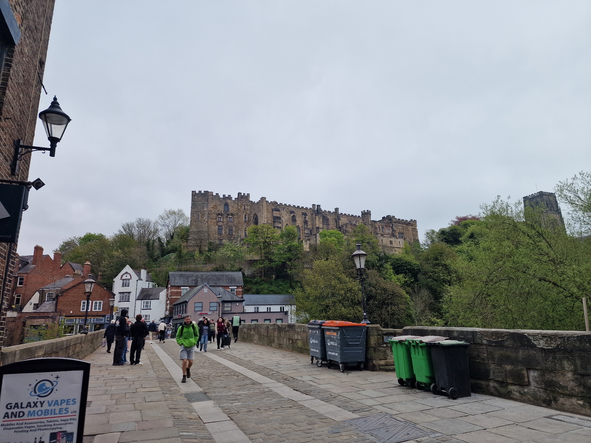 The image shows a scene from the city of Durham, United Kingdom, with Durham Cathedral and Durham Castle visible in the background. The view is likely from Framwellgate Bridge looking towards the cathedral. The foreground shows a cobblestone bridge with people walking in both directions. A man in a green jacket and shorts walks towards the camera. On the right side of the bridge are a row of several waste bins, and on the left is a sign advertising "Galaxy Vapes and Mobiles LTD". The sky is overcast. The castle and cathedral are situated on a hill covered with trees. Several buildings are visible below the castle. A lamppost is visible on the left and right side of the bridge.