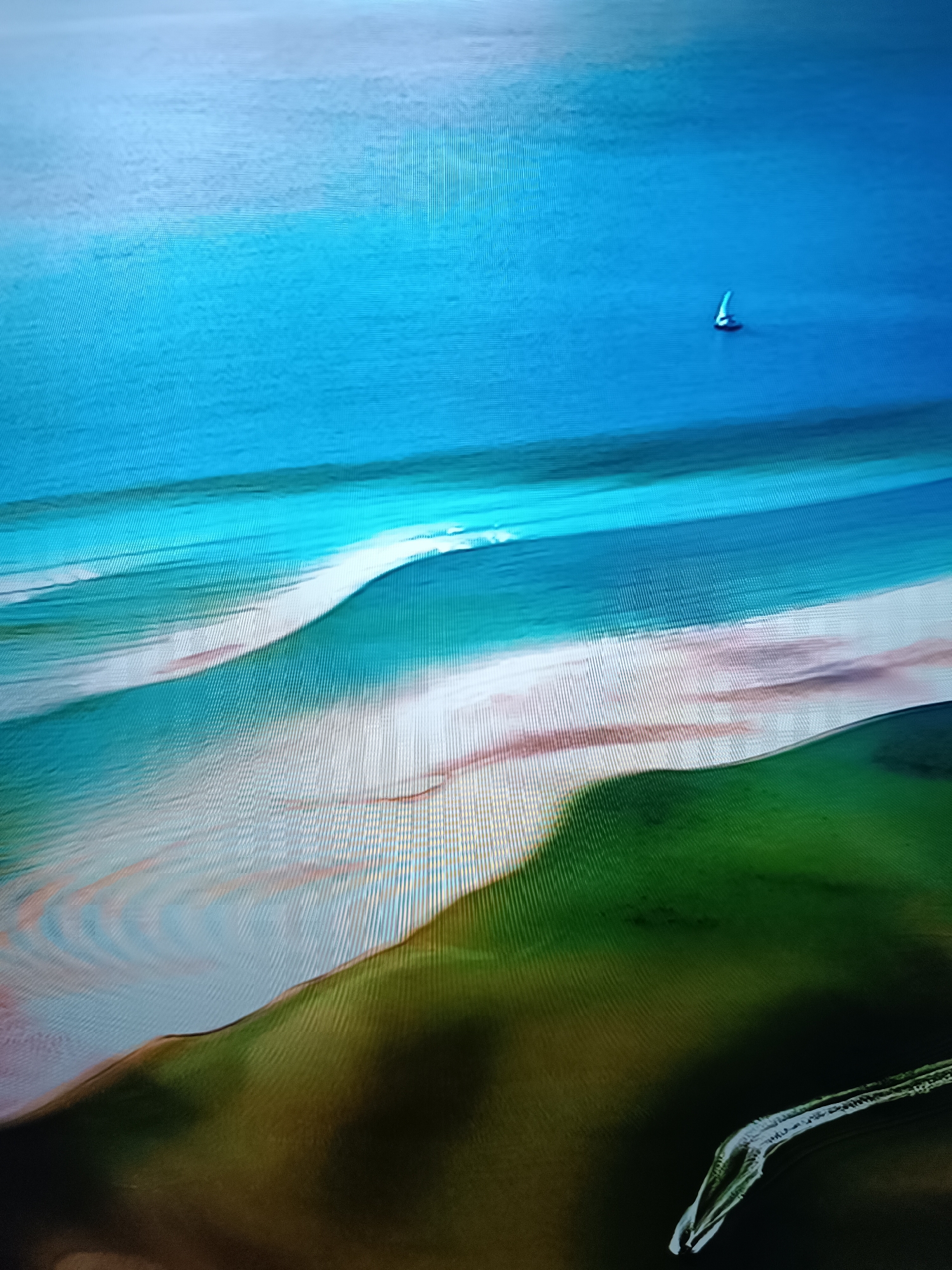 An aerial view shows a serene coastal landscape. The image features a vast expanse of calm blue water, transitioning into lighter turquoise hues closer to the shore. A small white sailboat with a single sail is visible in the distance, glinting against the deep blue sea. The coastline is characterized by sandy stretches and vibrant green land, suggesting a natural, undeveloped area. The composition is dominated by broad strokes of color, with the water occupying the upper half of the frame and the landmasses filling the bottom. The overall impression is one of tranquility and natural beauty. The image does not depict any urban elements or specific landmarks that would allow for identification of the city of Taraz city administration, Kazakhstan.