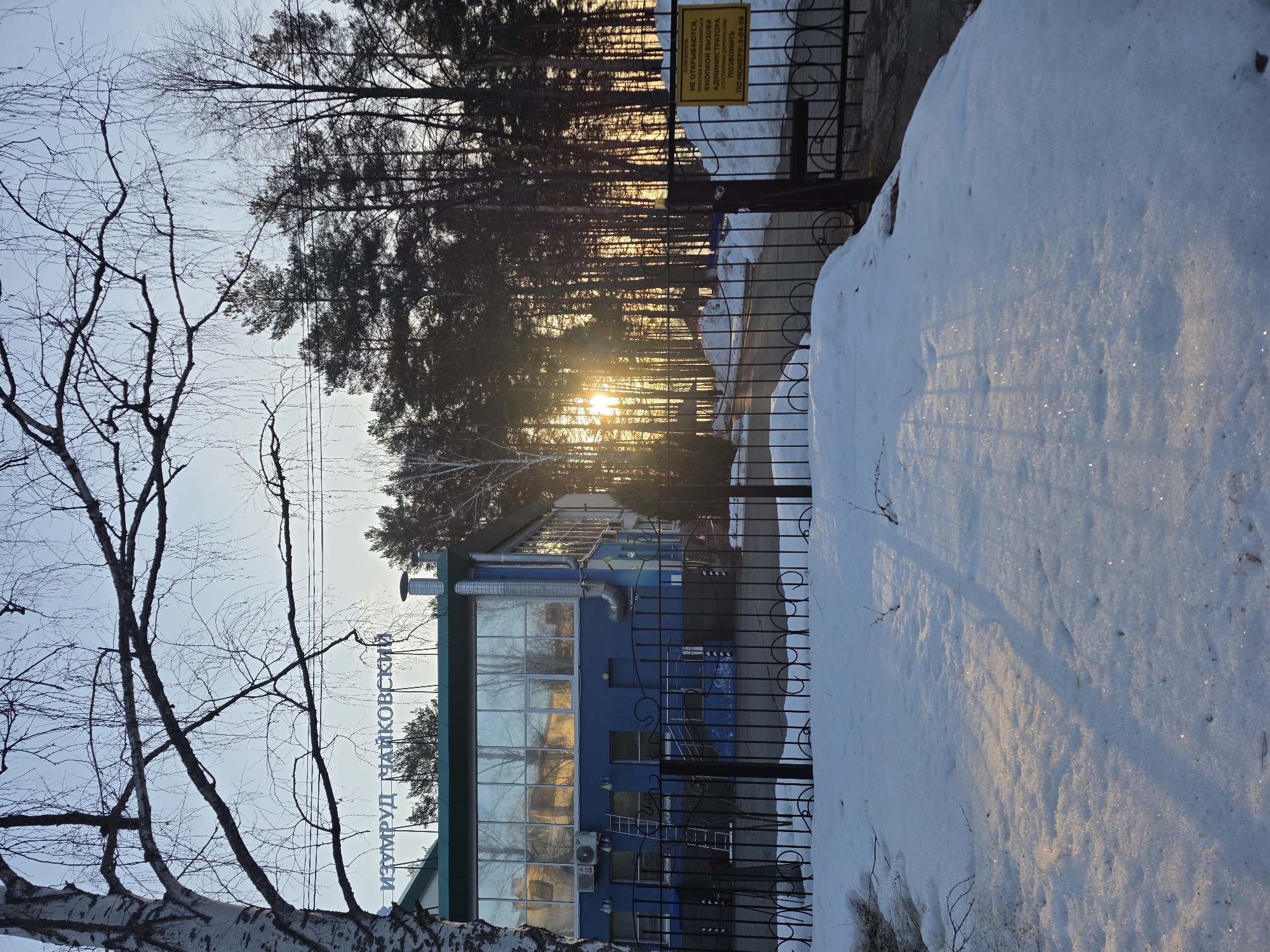 The image depicts a building in the city of Chaikovsky, Russia, with the name "IZUMRUD" displayed in Cyrillic script. The building has a blue facade with large glass windows reflecting the surrounding environment. A snowdrift is visible in the foreground along a wrought iron fence. Trees are in the background with the sun shining through them. A yellow sign with Cyrillic text is attached to the fence. A bare birch tree is visible.