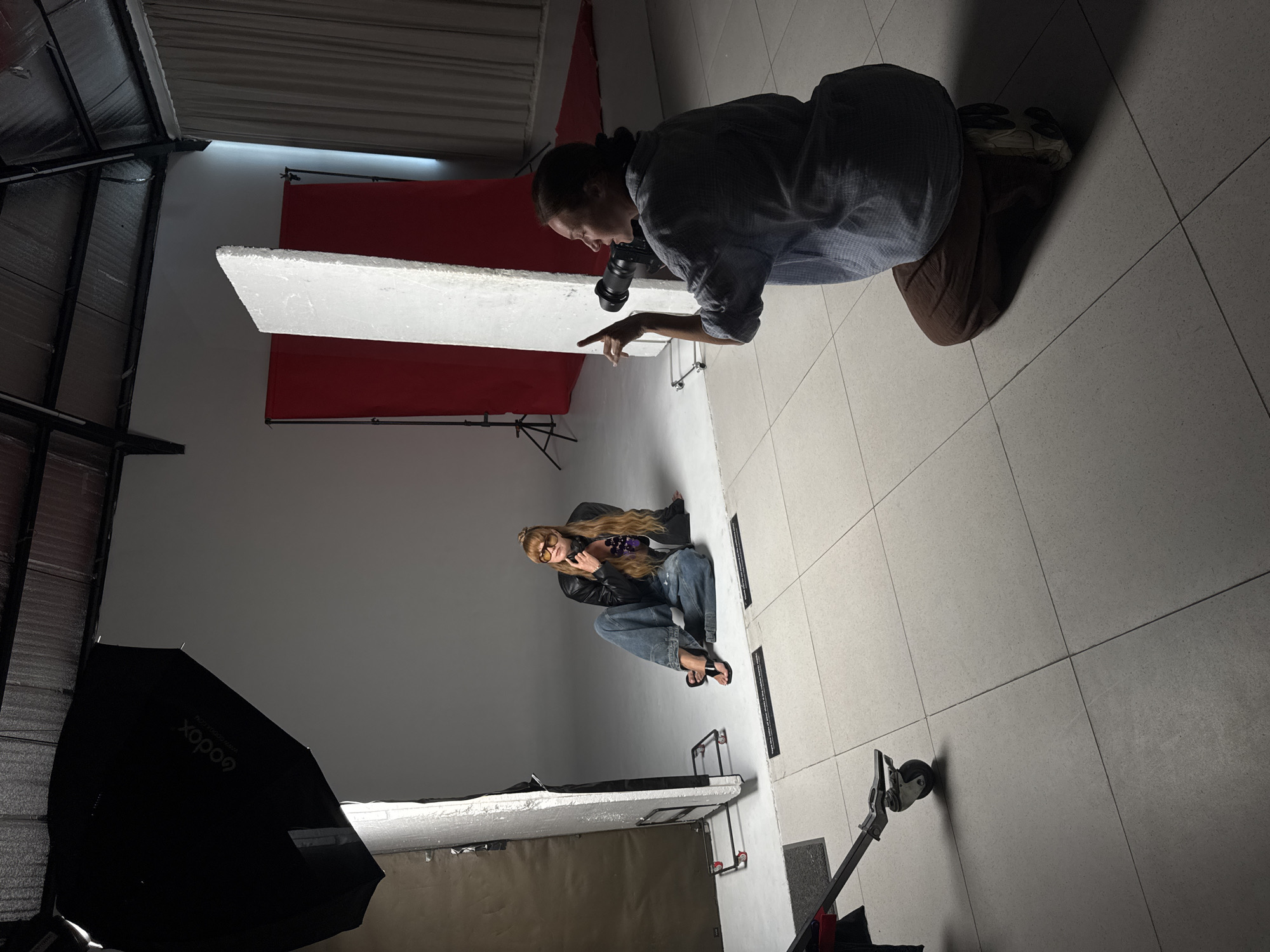 This media file captures a photoshoot in an indoor studio setting in Denpasar Barat, Indonesia. The scene features two individuals: a photographer and a model, along with various pieces of photography equipment.
In the foreground on the right, a person identified as the photographer is kneeling on a light grey tiled floor. They wear a dark plaid shirt, brown pants, and sneakers. They are holding a black camera and appear to be giving directions or pointing towards the model with their right index finger. Their dark hair is tied back.
Towards the left, another person, the model, is sitting in a crouched position on what appears to be a light grey wall (which, when the image is oriented correctly, is the studio floor). The model has long, light-colored hair and is wearing a black leather jacket, baggy blue jeans, and black open-toed sandals. They are also wearing orange-tinted glasses and holding a dark object in their hands, possibly a prop.
The studio environment includes a vibrant red fabric backdrop visible behind the photographer. A large white reflector board stands vertically between the two individuals. To the far left, a black photography umbrella or softbox is partially visible, with the brand name "Godox" printed on it in white text. Other studio equipment, such as metal stands and a wheeled light stand, are also present on the tiled floor. The lighting is artificial, characteristic of a photography studio, casting soft shadows.
The original image is rotated 90 degrees clockwise from a standard upright orientation. The overall activity depicted is a professional photography session.