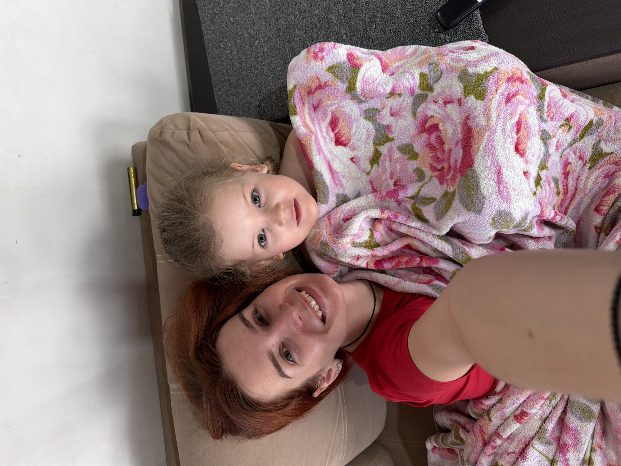 The image displays an adult female and a young female child in an indoor setting within the city of Chaikovsky, Russia. The adult female, positioned on the left, has reddish-brown hair, a light complexion, and is wearing a red short-sleeved top and a black choker. She is smiling broadly and holding the camera for a selfie. The child, situated on the right, has light brown hair and a light complexion, and is partially wrapped in a plush blanket adorned with a pink, white, and green floral pattern. Both individuals are lying on a light brown upholstered surface, likely a sofa or bed, and are looking directly at the camera. In the upper right quadrant, behind the child, a gold-colored cylindrical object and a purple item are visible on the upholstery. The background features a plain light-colored wall at the top and a dark gray carpet or rug along the right side. A dark rectangular object is partially visible on the carpet in the bottom right corner.