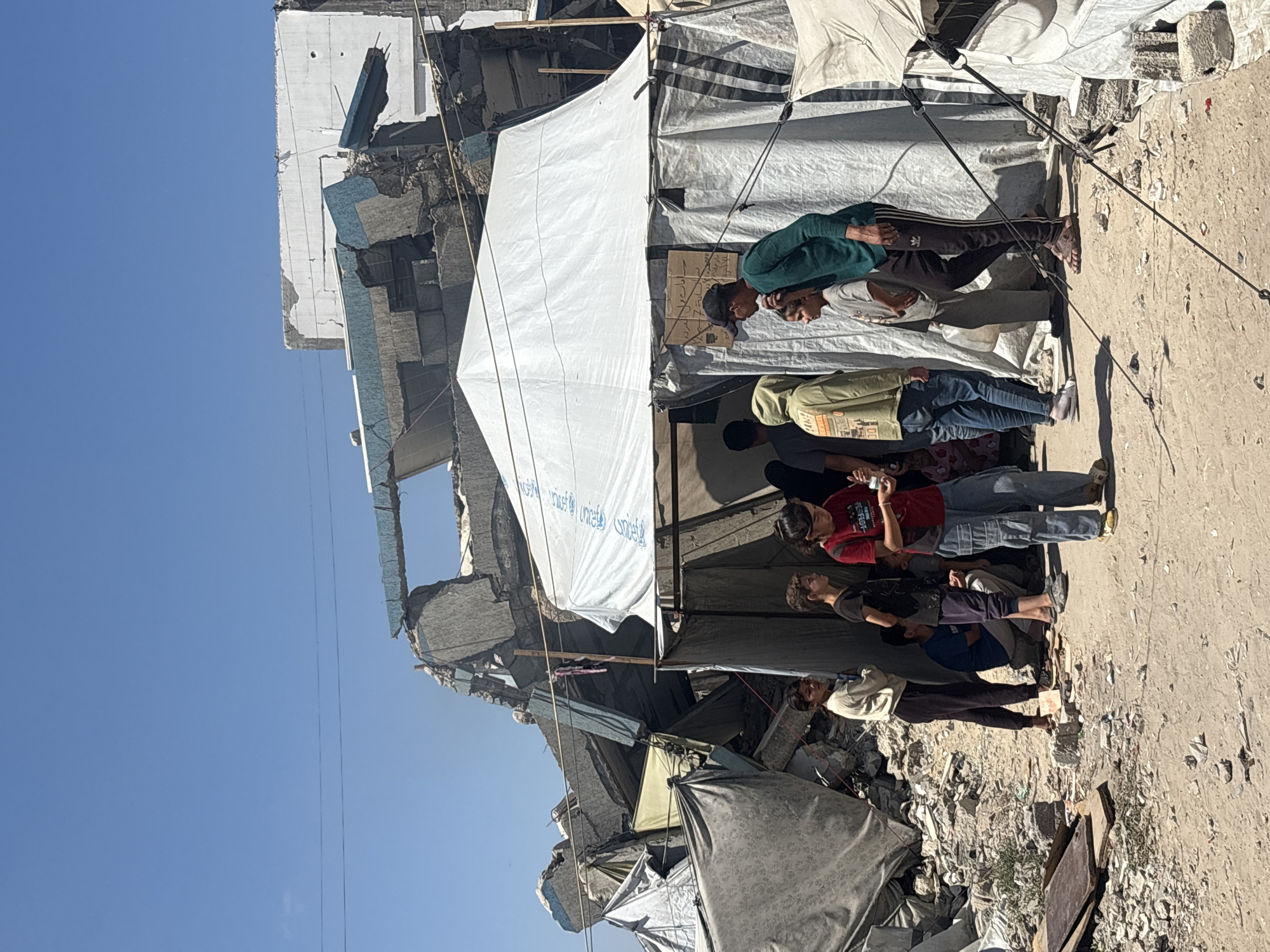 The image depicts a group of people standing near a partially destroyed building under a clear blue sky. The building appears to be made of concrete and has large sections missing or collapsed, revealing exposed rebar and rubble. Several temporary shelters, likely made of tarps and fabric, are erected in front of the damaged structure.
The people are varied in age and gender, dressed in casual clothing. Some are looking at their phones, while others appear to be in conversation or observing the surroundings. The overall scene suggests a sense of displacement or the aftermath of a destructive event, possibly an air strike or conflict, given the context of Gaza.
The time of day appears to be late morning or early afternoon, as indicated by the bright sunlight and the shadows cast on the ground. The weather is sunny and clear. The ground is dusty and littered with debris, consistent with a damaged urban environment.
One visible detail is a white tarp with repeating text, which appears to be the UNICEF logo and text in Arabic. The emotions conveyed by the people are difficult to ascertain definitively, but the context of the scene might suggest resilience, coping, or a somber mood.