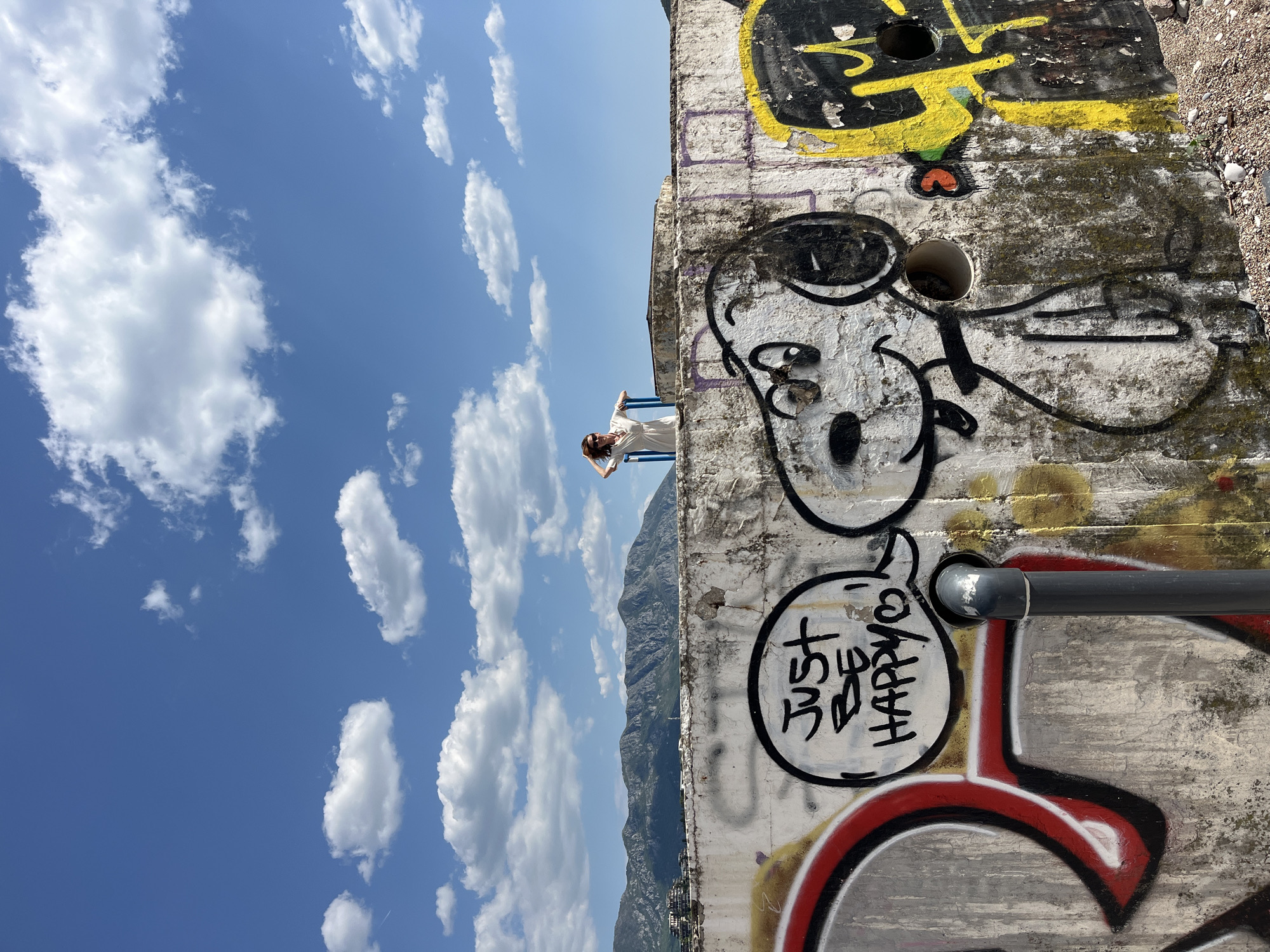 A woman in a white dress stands on a blue railing in Budva, Montenegro. She is in front of a concrete wall covered in graffiti, including cartoonish figures and text that reads "Just be happy." The background shows a blue sky with scattered clouds and mountains in the distance.
