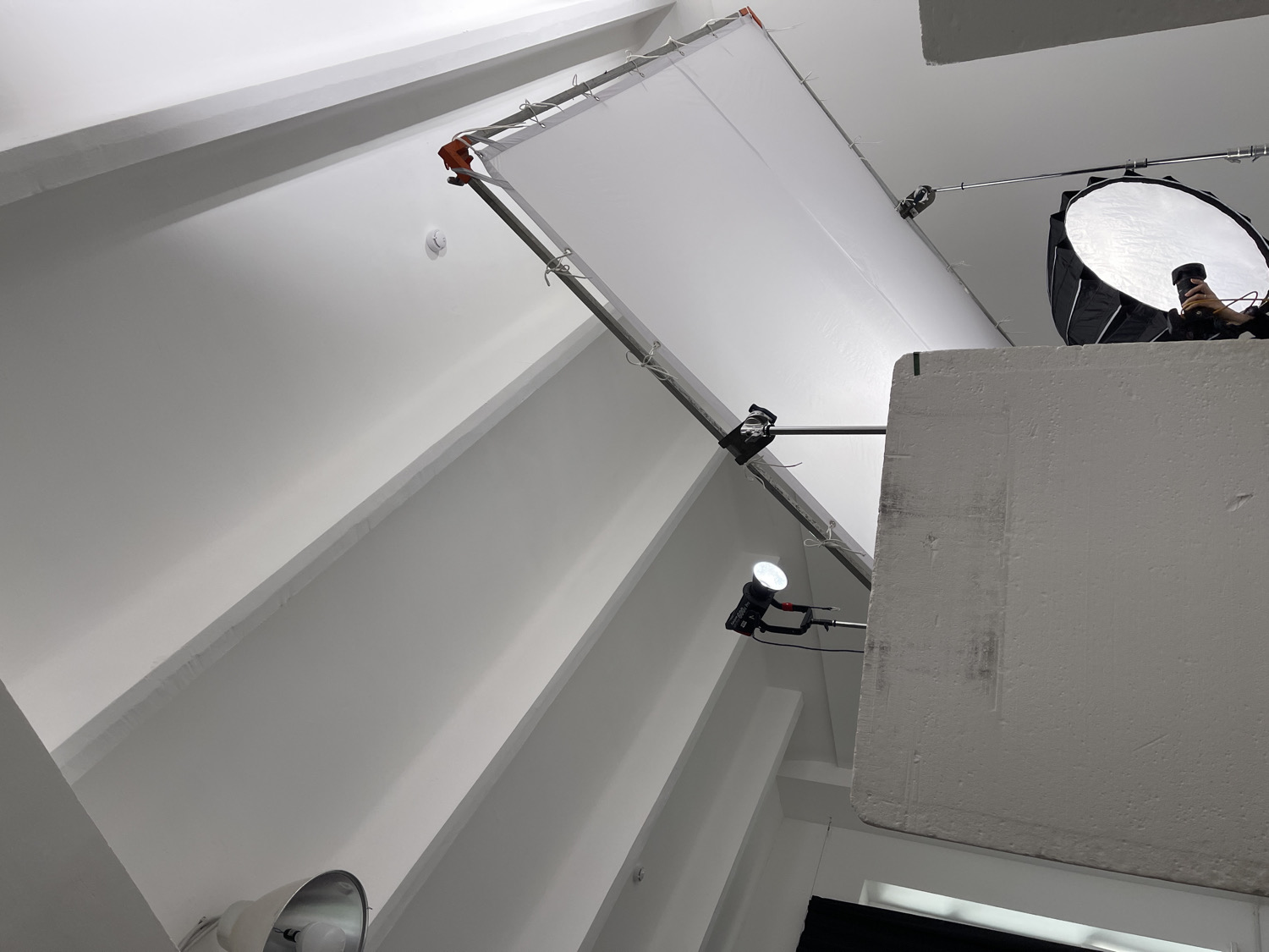 The image provides an upward view into what appears to be a photography or film studio, characterized by predominantly white walls and ceiling. The setting is brightly illuminated by artificial lighting equipment.
A large, rectangular white fabric diffuser or scrim is suspended from the ceiling in the upper left and central portions of the frame, supported by metal rods and clamps. To its right, a circular softbox or beauty dish with a silver reflective interior is visible, accompanied by a hand holding a camera, suggesting an active photography or videography session.
In the mid-right, a thick, white rectangular foam board, likely used as a reflector or flag, stands vertically. Further to the left and below the main diffuser, a small, bright LED light or flash unit is mounted on an arm, directing its beam upwards. Another light fixture is partially visible in the lower left.
The composition emphasizes the geometric lines of the studio's interior, with white structural beams or elements on the ceiling. Various clamps, rods, and stands are used to position the lighting modifiers. The scene is devoid of people apart from the hand holding the camera, and no discernible text is visible. The controlled indoor lighting makes the time of day indeterminate.