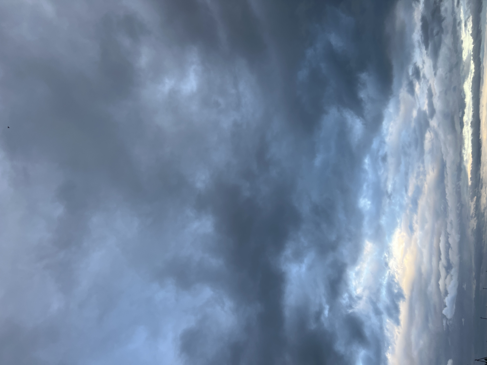 The image shows a dramatic sky filled with dark, heavy clouds, suggesting an impending storm. The clouds are dense and textured, with varying shades of gray and blue, hinting at atmospheric depth. In the far upper center of the image, a tiny, dark silhouette, possibly a bird or a distant aircraft, is visible against the overcast sky. The lower portion of the image reveals a sliver of lighter sky, with hints of yellow and pale blue, suggesting the presence of the sun attempting to break through or the approaching dawn or dusk. The scene is devoid of any specific landmarks or elements that would tie it to Калодзежскі сельскі Савет, Belarus, or any other urban location. The focus is entirely on the atmospheric conditions of the sky.