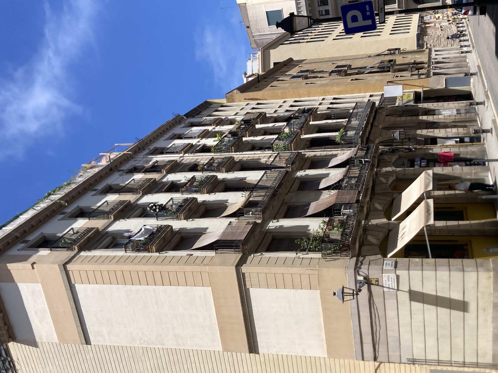 The image shows a street scene in Barcelona, Spain, on a bright, sunny day with a clear blue sky. The foreground features a multi-story building with a facade that is a mix of light cream and a soft beige. The building has numerous windows with black wrought-iron balconies, many adorned with potted plants and some with rolled-up awnings. Some balconies have clothes hanging to dry.
To the left of the main building, a portion of another building with horizontal beige siding is visible. A street lamp is mounted on the corner of the cream and beige building. Below the balconies, there are shops with awnings. One shop has a yellow facade and a sign that is not clearly legible. Another shop has a white sign.
Further down the street, other buildings with varied architectural styles and colors are visible, including a tall, yellowish building and a lighter-colored building in the background. A black lamppost is positioned on the right side of the street. A blue parking sign with the letter "P" and "20 min" is also visible on the right.
There are a few people visible: one man standing near a shop entrance on the left, and two women walking further down the street on the right. A car is partially visible in the lower right corner of the image. The street is paved with cobblestones and has yellow lines marking the road. The overall impression is of a vibrant urban environment in Barcelona, likely in a historic district, with a mix of residential and commercial spaces.