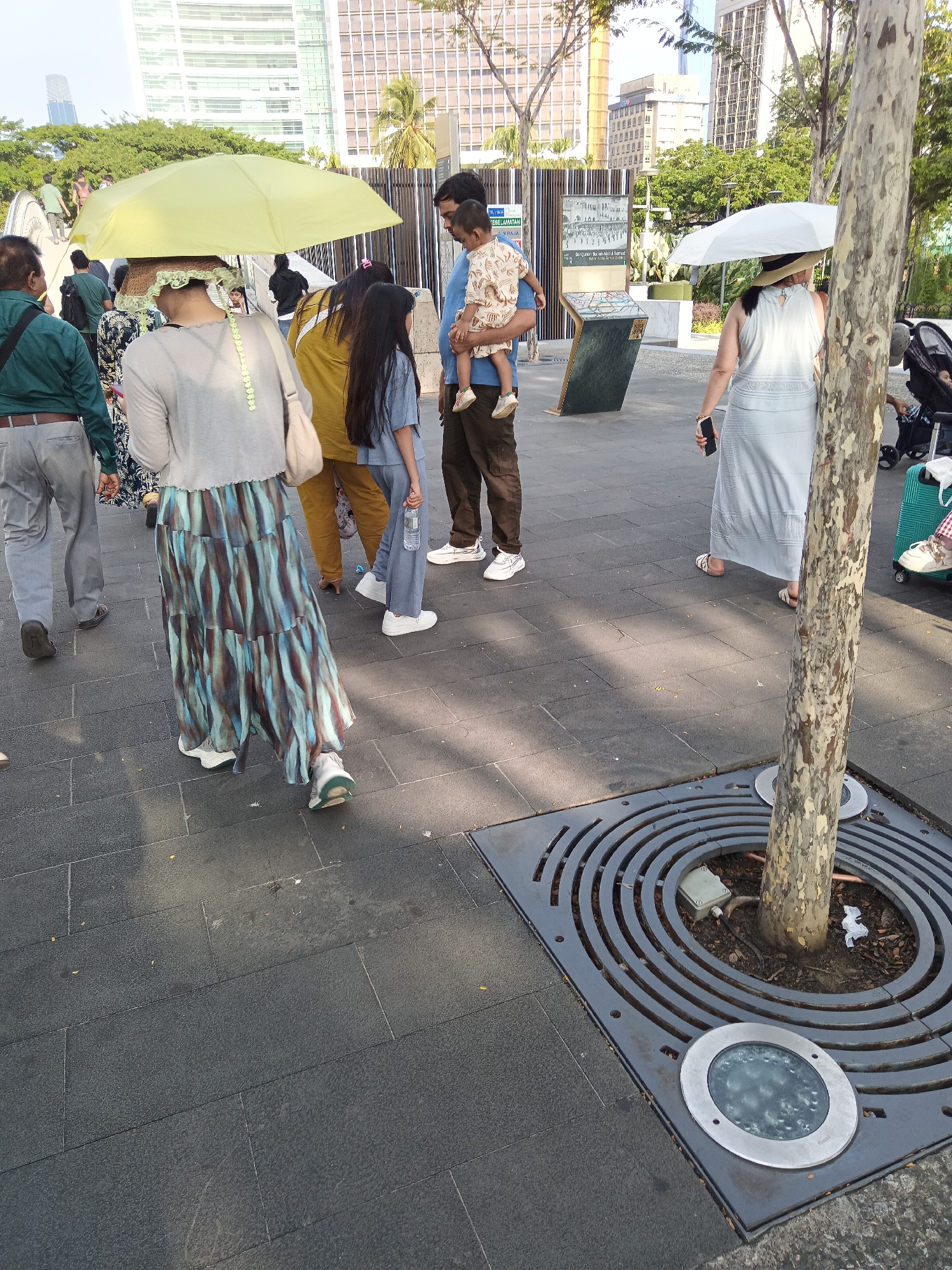 In this image, a diverse group of people is captured walking on a paved walkway in an urban park. The scene is bathed in natural sunlight, casting shadows on the ground. In the foreground, a woman is walking away from the camera, her body obscured by a flowing, multi-colored skirt and a light-colored shirt. She is carrying a handbag over her shoulder. A large, bright yellow umbrella is held above her, offering shade.
To her left, a man in a green shirt and gray pants is walking towards the camera. A man in a blue shirt is seen carrying a young child, who is wearing a patterned outfit. Beside them, a young girl in a blue outfit is walking, holding a clear plastic bottle.
In the middle ground, another woman is walking away from the camera, holding a white umbrella and a smartphone. She is wearing a light-colored dress and sandals. Behind her, a stroller with a baby is visible. The background reveals modern buildings and lush green trees, indicating an urban environment. The paved walkway is made of dark rectangular tiles. A circular metal grate surrounds a tree in the foreground, with embedded lights illuminating the ground. The presence of modern architecture and trees suggests this location is within Kuala Lumpur, Malaysia, potentially in a public park or recreational area.