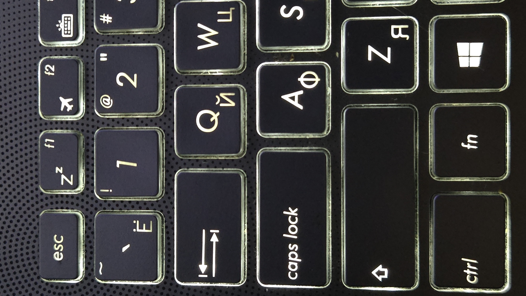 This is a close-up, high-angle shot of a black laptop keyboard. The keys are illuminated from beneath with a white light, creating a glow around their edges. The keys have both Latin and Cyrillic characters on them. Visible keys include "esc", "f1", "f2", "~", "Ё", "!", "1", "@", "2", "#", "3", a symbol resembling two arrows pointing away from each other, "Q", "Й", "W", "Ц", "caps lock", "A", "Ф", "S", a large blank key with an upward arrow symbol, "Z", "Я", "ctrl", "fn", and a Windows logo key. The background is a dark, textured surface with a dotted pattern, likely the palm rest of the laptop. The image provides no information about the city of Lebedyan, Russia, or any other specific location.