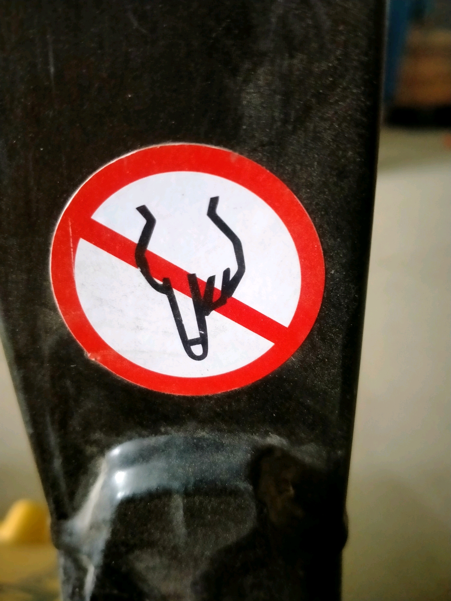 A circular sticker with a red border and a red diagonal line across it is affixed to a black surface. Inside the circle, a stylized black image depicts a phallus with a thumb and forefinger extended. The sticker is positioned on what appears to be part of a machine or equipment. The background is dark and out of focus, with a hint of yellow and gray to the left and right. The sticker's context within the city of Klepu, Indonesia, is not discernable from the image itself.