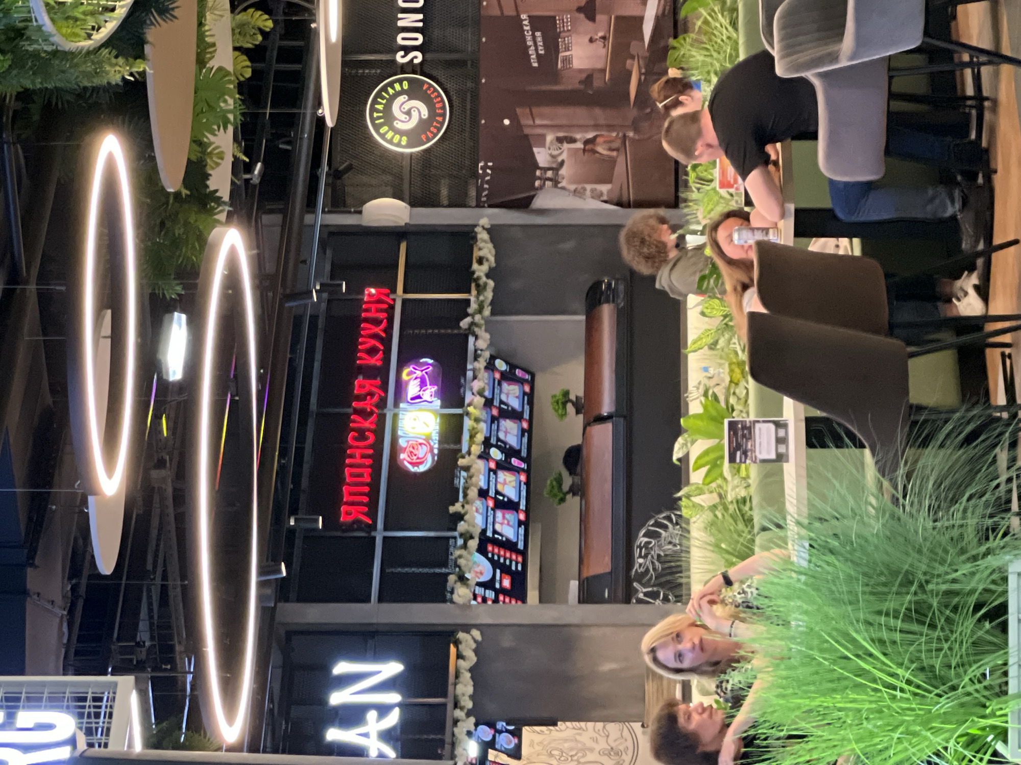 This image depicts the interior of a modern food court or restaurant area. In the foreground, several people are seated at tables. On the left, a man and a woman with blonde hair are visible, with the woman looking away from the camera. In the center and right foreground, other individuals are seated, one of whom appears to be leaning on a table with a can of soda. Green foliage is prominent throughout the scene, suggesting an attempt to bring nature indoors.
Above the diners, there are prominent circular light fixtures that emit a bright, white light. Behind the seating area, a counter or display area is visible with menus or pictures of food items. Signs with Russian text are also present, including one that reads "ЯПОНСКАЯ КУХНЯ" (Japanese Cuisine) in red neon letters. Another sign features a circular logo with the text "SONO ITALIANO PASTA FRESCA" and the text "SONO" in large letters.
The overall impression is a contemporary dining establishment. The location, Ladozhskoe Ozero, Russia, is an area on the eastern side of Saint Petersburg, known for its residential developments and commercial centers. This scene is likely within one of these commercial centers. The architecture and decor suggest a relatively new or recently renovated space.