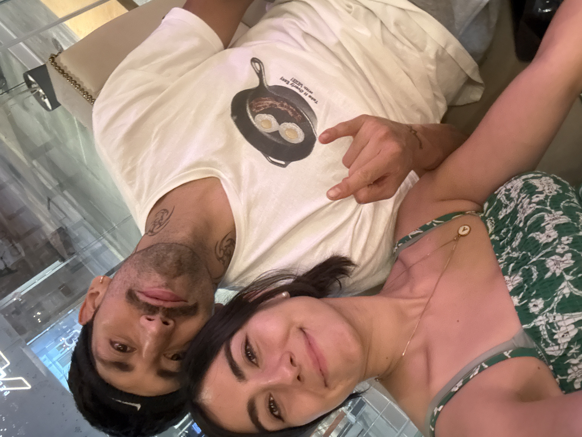 A photograph taken indoors in the city of Puebla, Mexico, captures a man and a woman lying together, presumably taking a selfie. The man has a mustache, a thin gold earring in his left ear, and dark hair. He is wearing a white t-shirt with a graphic print of eggs in a skillet. His right hand, with a visible wrist tattoo, is pointing towards the graphic on his shirt. The woman is wearing a green and white floral-patterned dress, with a thin gold necklace. She has dark hair and is wearing a small, white pearl earring. The background suggests a modern, urban environment with glass surfaces and metallic accents.