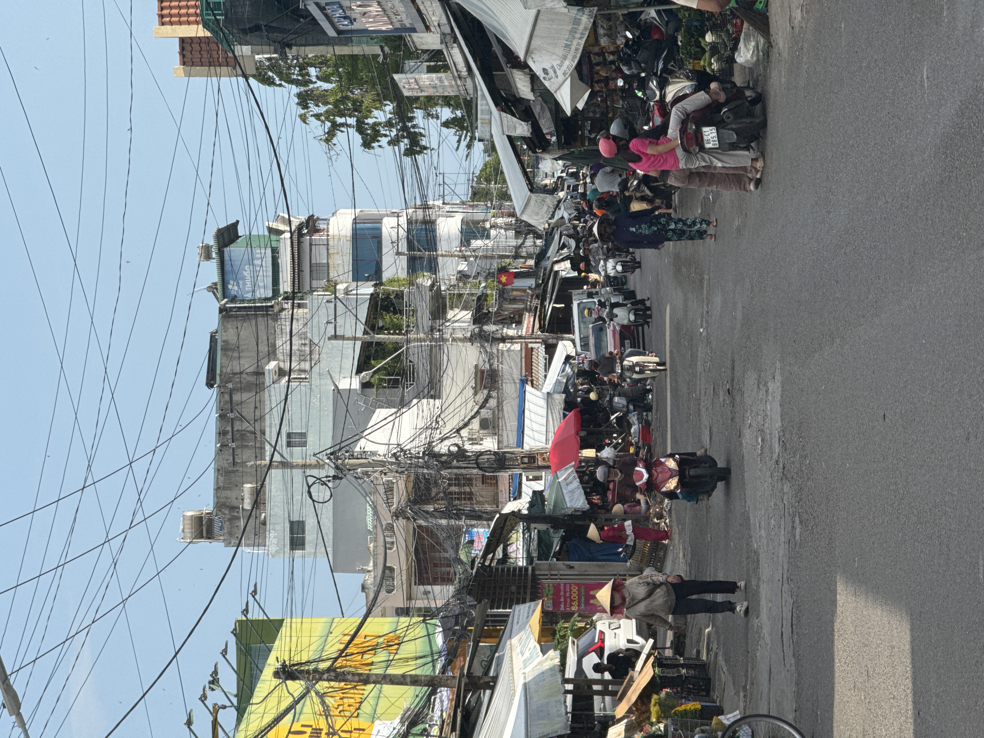 The image depicts a street scene in Phú Thủy, Vietnam, likely a market area, characterized by a mix of buildings, stalls, and a considerable amount of overhead electrical wiring. In the foreground, the asphalt road shows signs of wear and tear. Several people are present, mostly walking or riding motorbikes.
On the left side of the street, a woman wearing a conical hat and a grey jacket walks forward, accompanied by a white car parked nearby. A vendor stall is visible, displaying flowers. Further down the street, more stalls line the road, with awnings providing shade.
The center of the image shows a busy street with motorbikes navigating through the crowds. A woman in a red outfit and a conical hat is seen walking. A red umbrella is open above a stall. Buildings of various heights are visible in the background, some with visible balconies and windows. The sky is clear and blue, with a prominent network of electrical wires crisscrossing the frame.
On the right side of the street, more market stalls are set up under awnings, displaying goods, including what appear to be produce like watermelons. Several motorbikes are parked and in use. People are seen browsing the stalls and interacting. One stall has signage with Vietnamese text and pricing. The overall impression is that of a bustling, everyday urban environment.