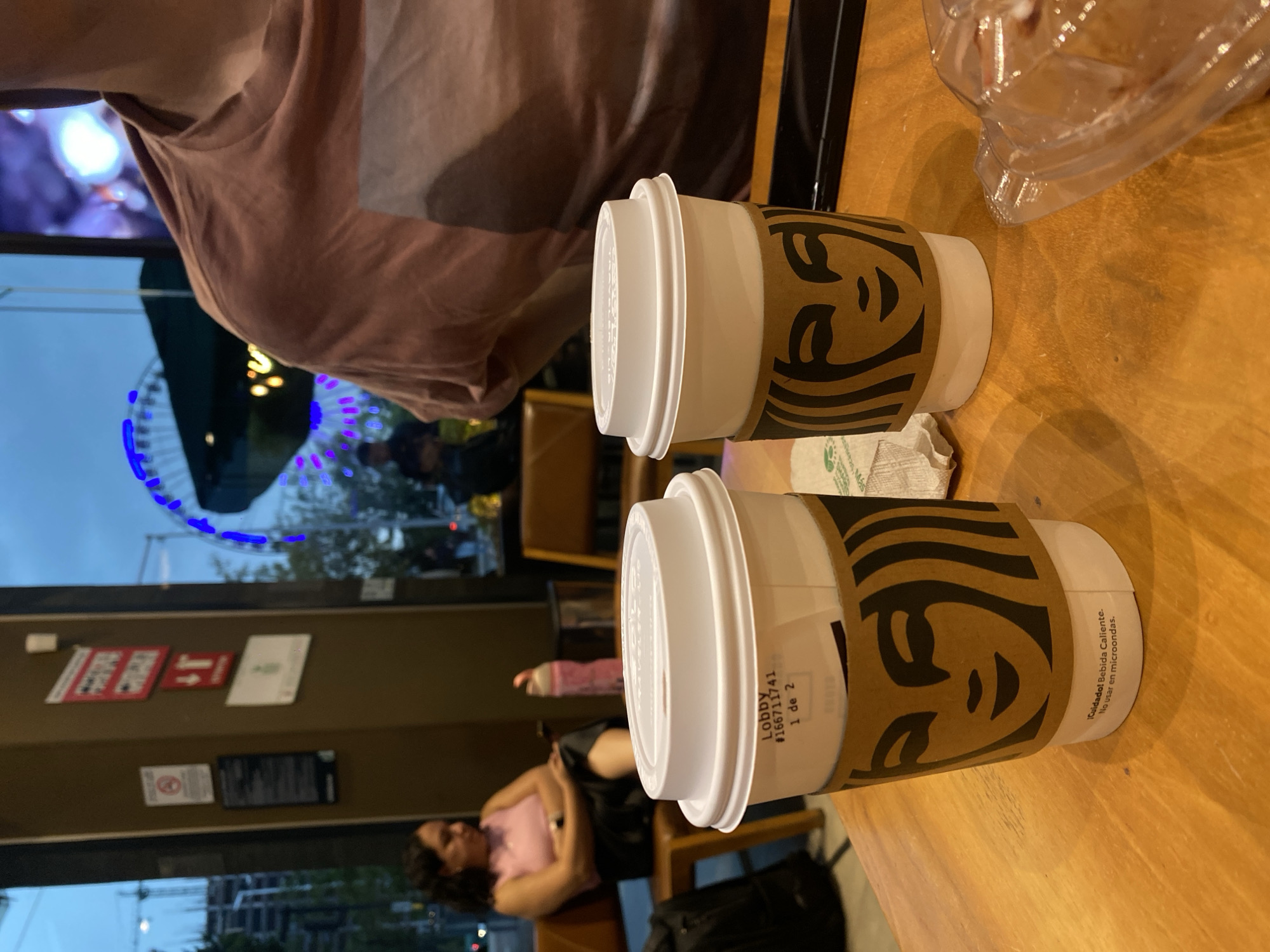 The image depicts a scene inside a Starbucks coffee shop. Two Starbucks coffee cups with lids are on a wooden table. One cup has a label that reads "Lobby #166711741 1 de 2 Cuidado! Bebida Caliente. No usar en microondas". In the background, through the glass window, one sees the "Estrella de Puebla" observation wheel, a notable landmark in Puebla, Mexico. A woman is reclining on a chair near the window. Signs and notices are visible on the interior wall behind the woman. Another person, partly visible, is seated at the table. A plastic food container is next to the coffee cups.